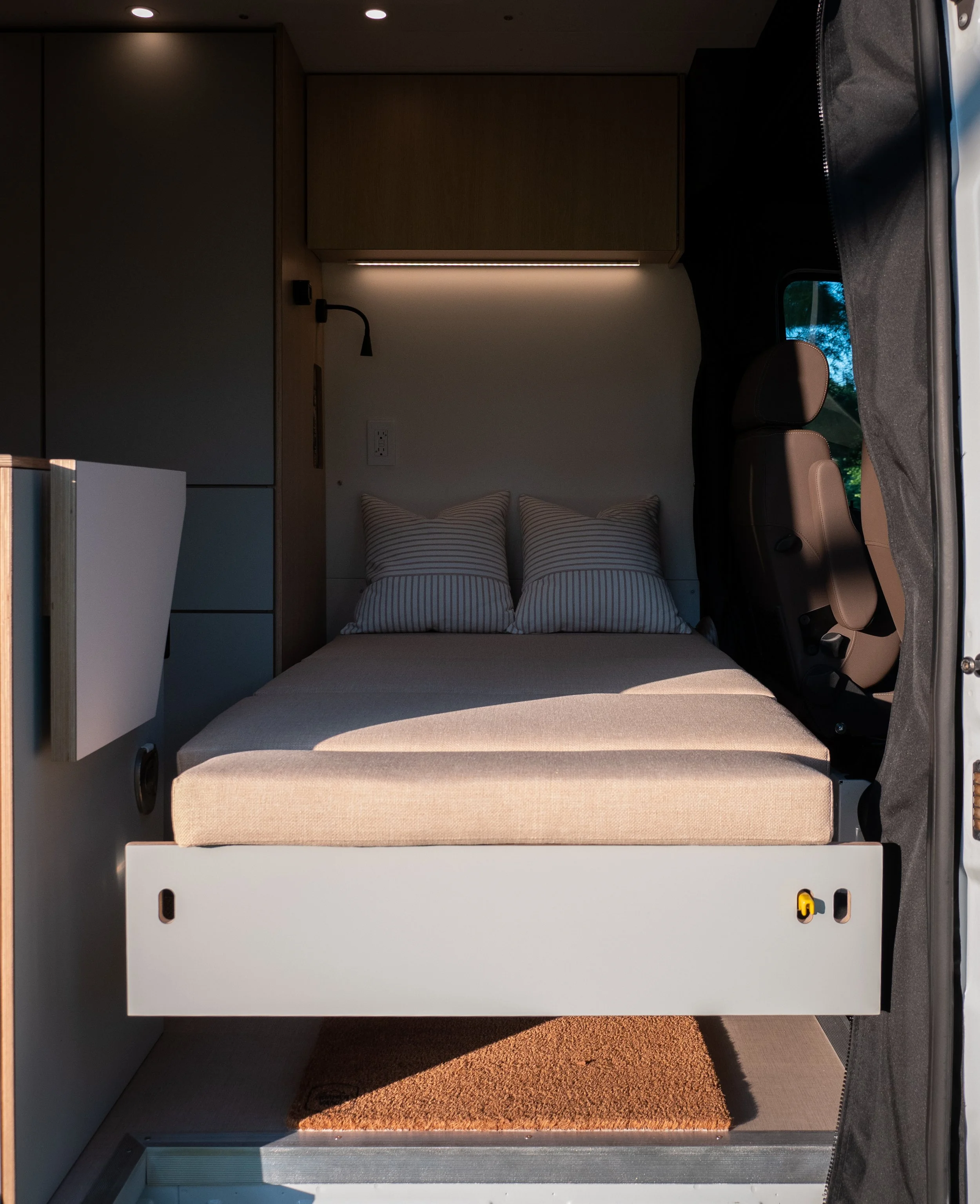 View through the open sliding door into 'Aina' Mercedes Sprinter van conversion — pull-out sleeping platform with beige cushion and two striped pillows, sage-green slide base with yellow latches; LED under-cabinet strip, black gooseneck reading sconc