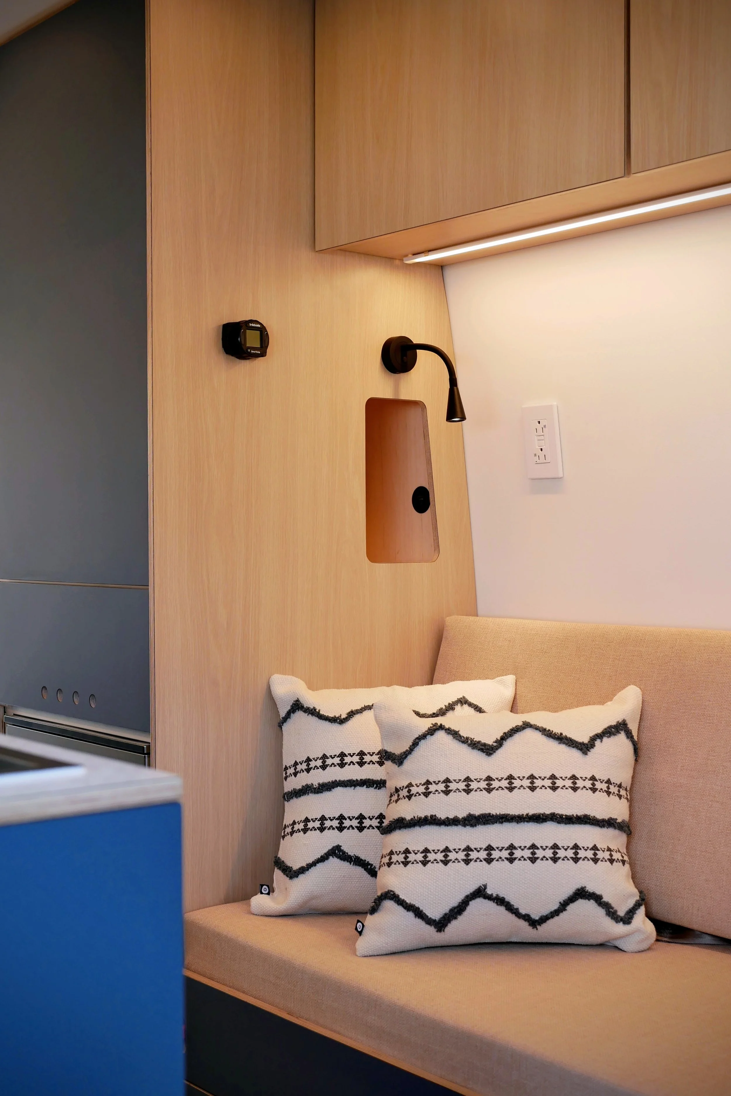 Lounge seating detail in 'Nordlys' — two geometric black-and-white cushions on cream bench seat, wall-mounted black reading lamp, recessed phone-charging nook in laminated birch plywood wall, LED strip lighting