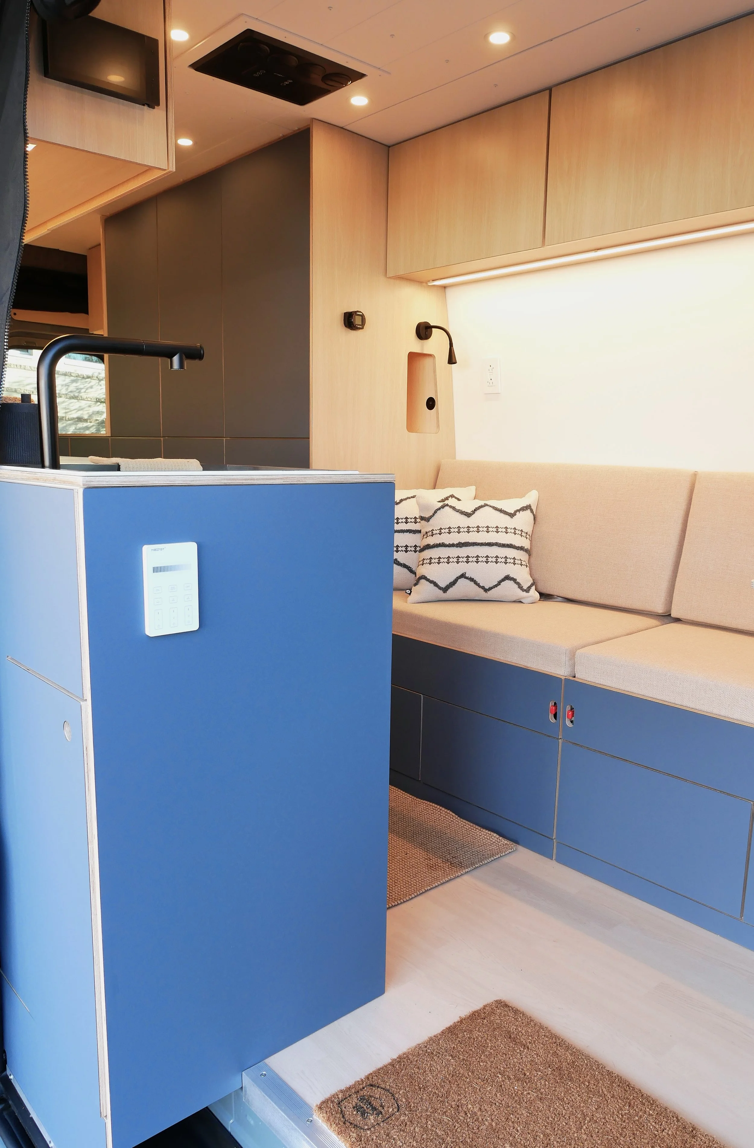 Interior overview of 'Nordlys' — blue kitchen counter with EcoFlow touch console in foreground, long beige bench seat along wall with geometric cushions, laminate ceiling with recessed lighting and 48V air conditioner
