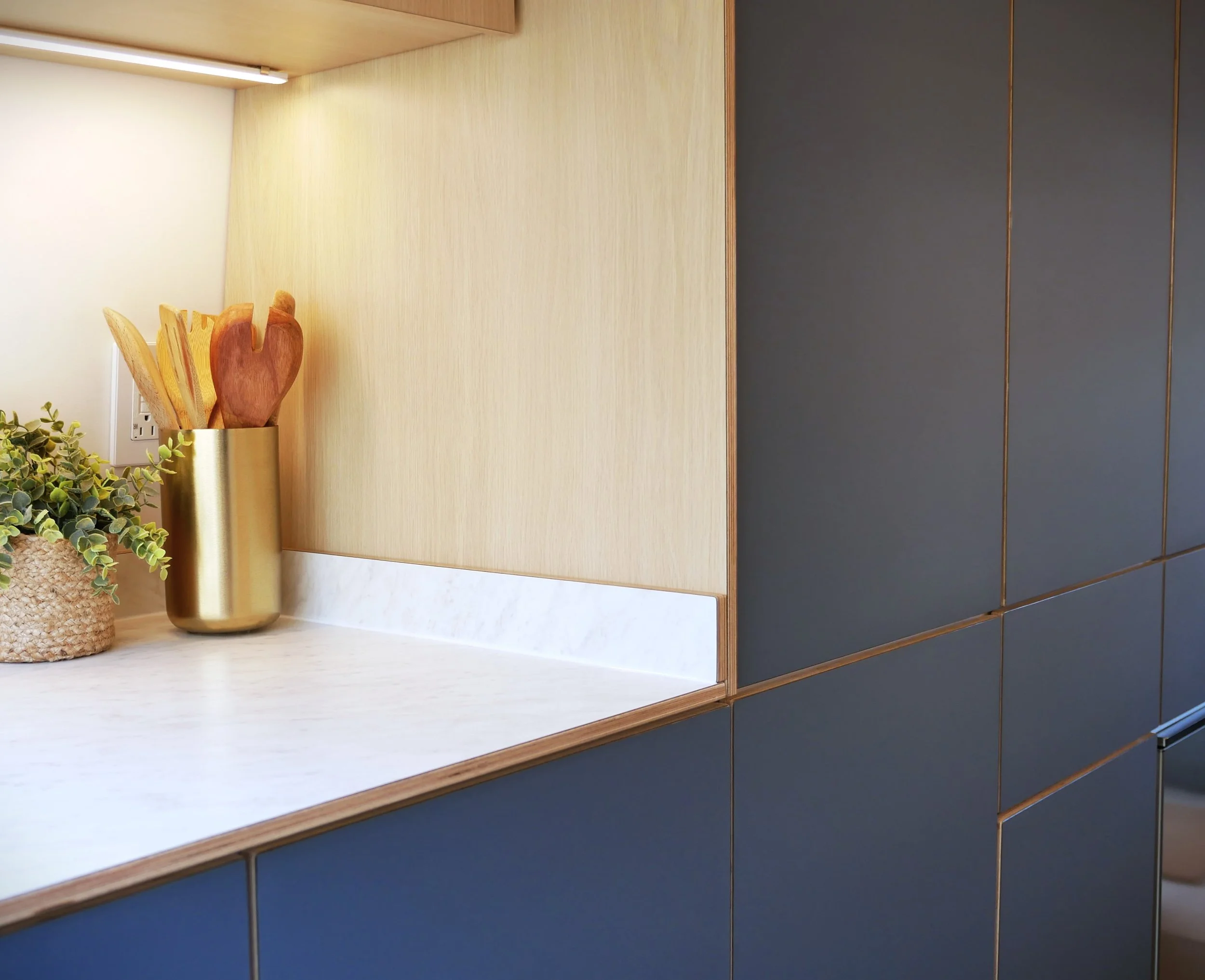 Kitchen backsplash in 'Nordlys' — plywood panel with LED strip, white countertop with plywood edge, brass utensil holder with wooden spoons, woven plant basket, blue cabinet doors