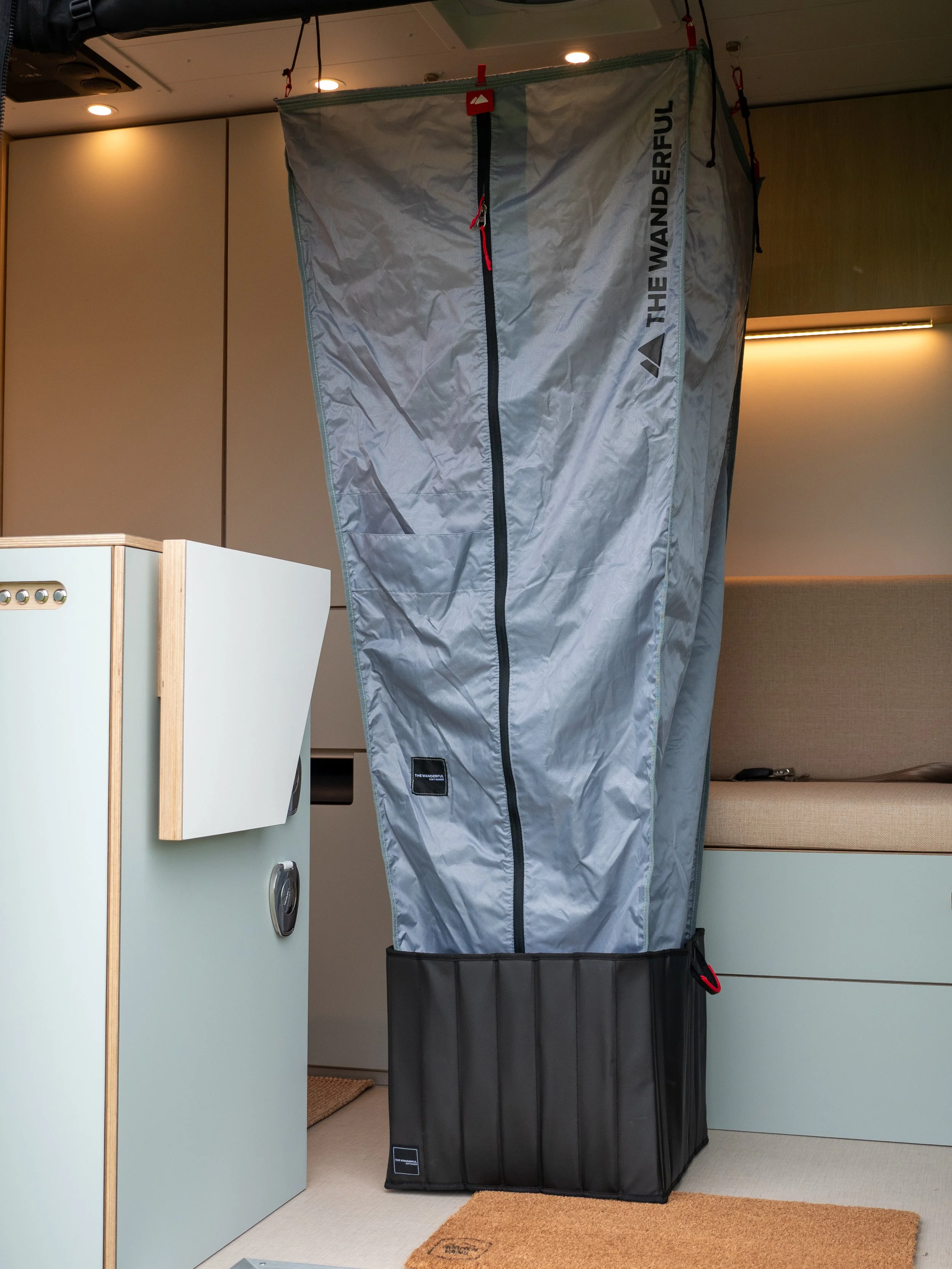 Portable shower setup inside 'Aina' Sprinter van conversion — grey waterproof pop-up shower tent with red zipper pulls and black base tray, suspended from ceiling hooks by red carabiners, sage-green cabinetry and linen bed platform visible behind it.
