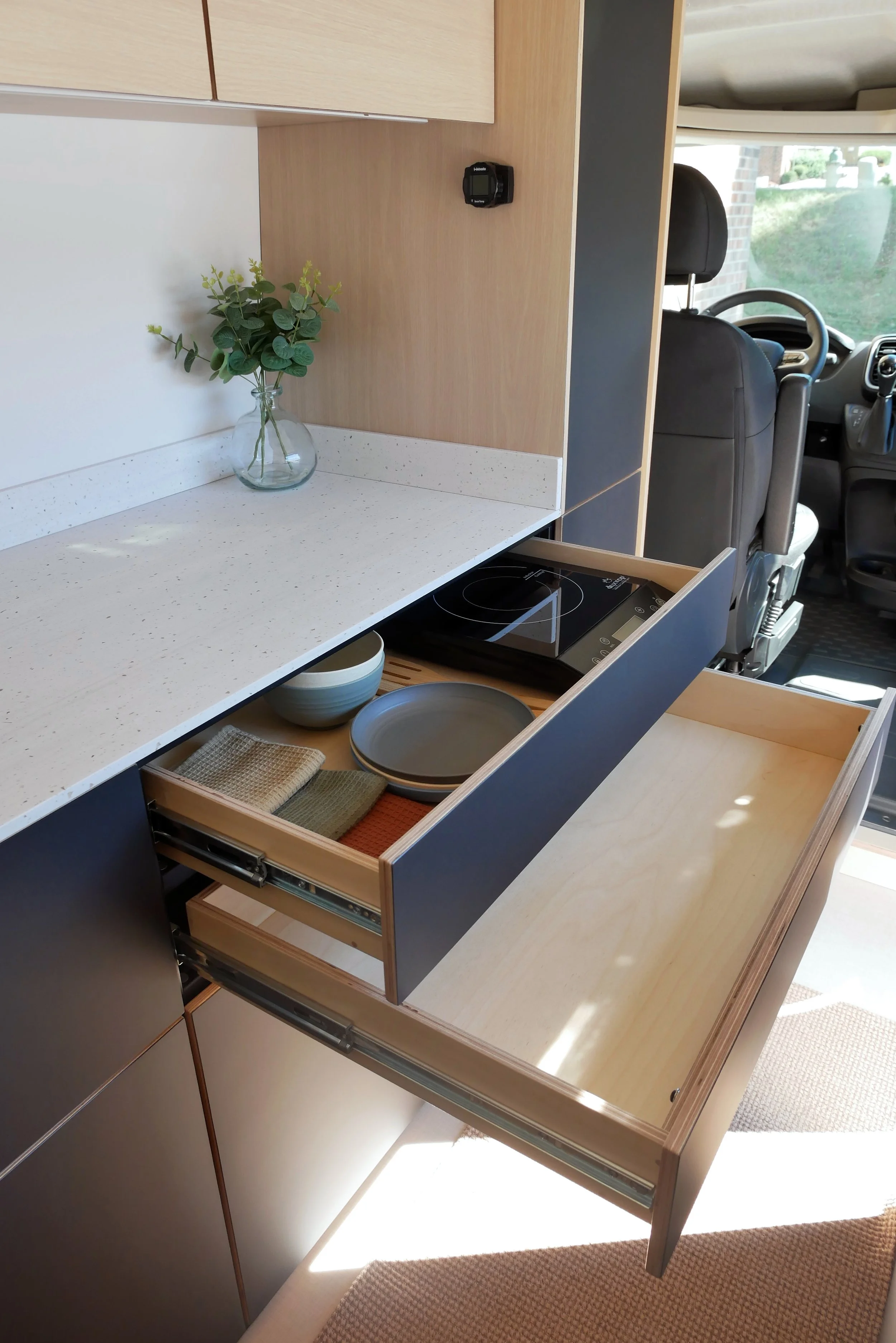 Van conversion galley kitchen in 'Bjorn' with two navy blue drawers fully extended — upper drawer reveals the hidden induction cooktop with bowls and plates; lower birch plywood drawer on heavy-duty slides; white quartz counter with eucalyptus vase a