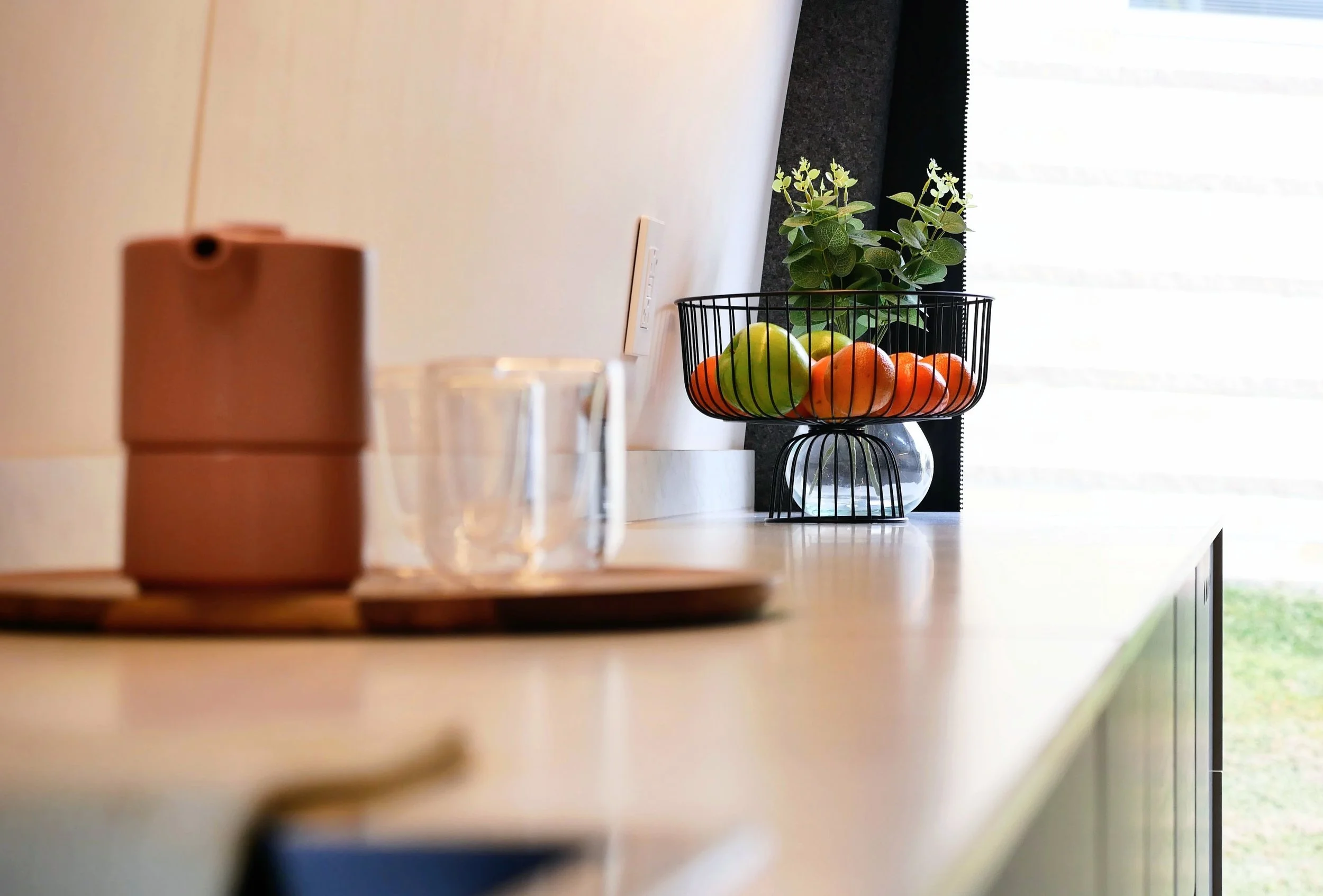 Kitchen counter lifestyle detail in 'Nordlys' — terracotta ceramic canister on wooden tray, glass cups, wire fruit bowl with fresh fruit, and small plant in natural window light
