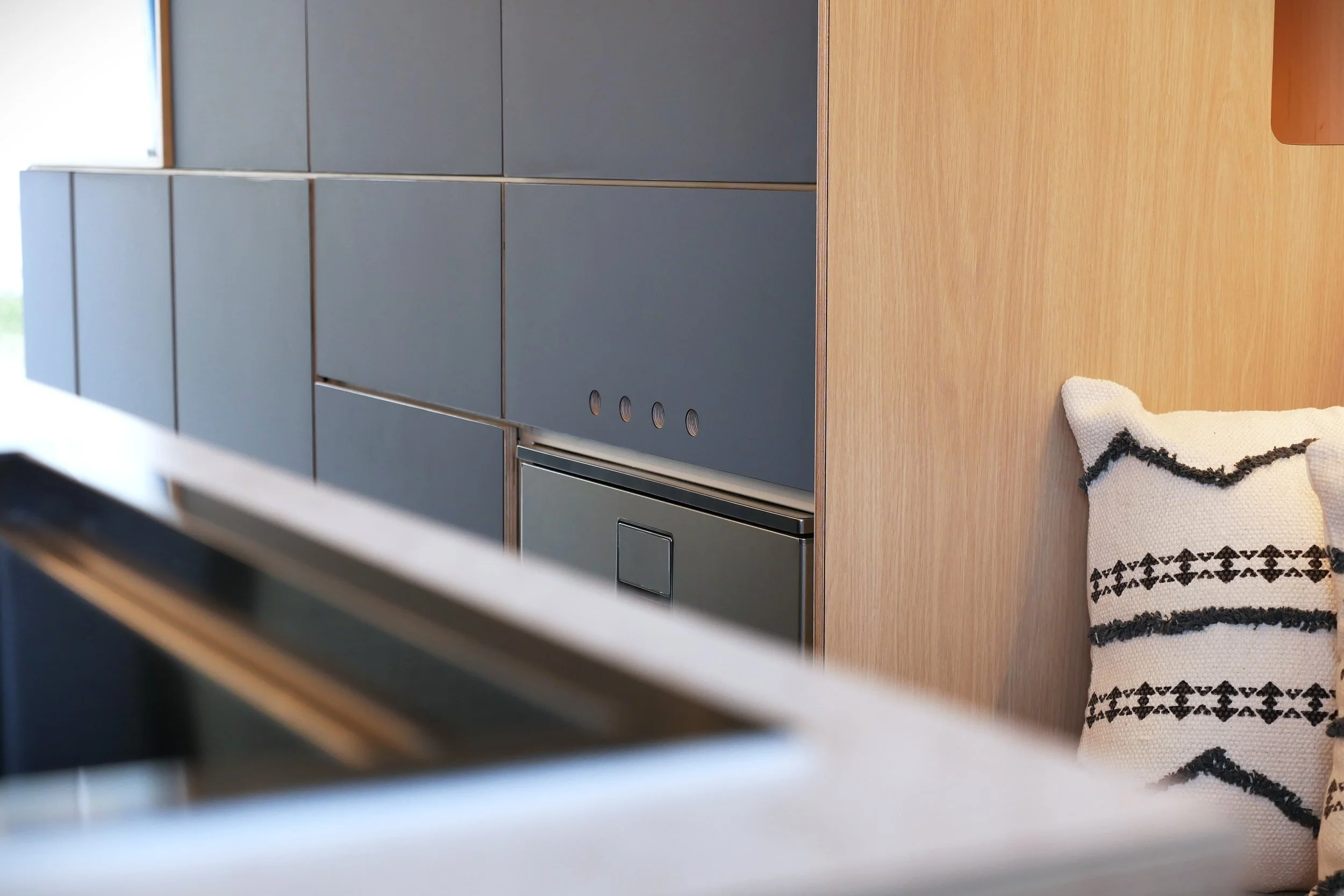 Cabinet detail in 'Nordlys' — dark blue laminate cabinet doors with plywood edges and integrated push-latch hardware, Indel drawer fridge and geometric cushion on bench seat beside them
