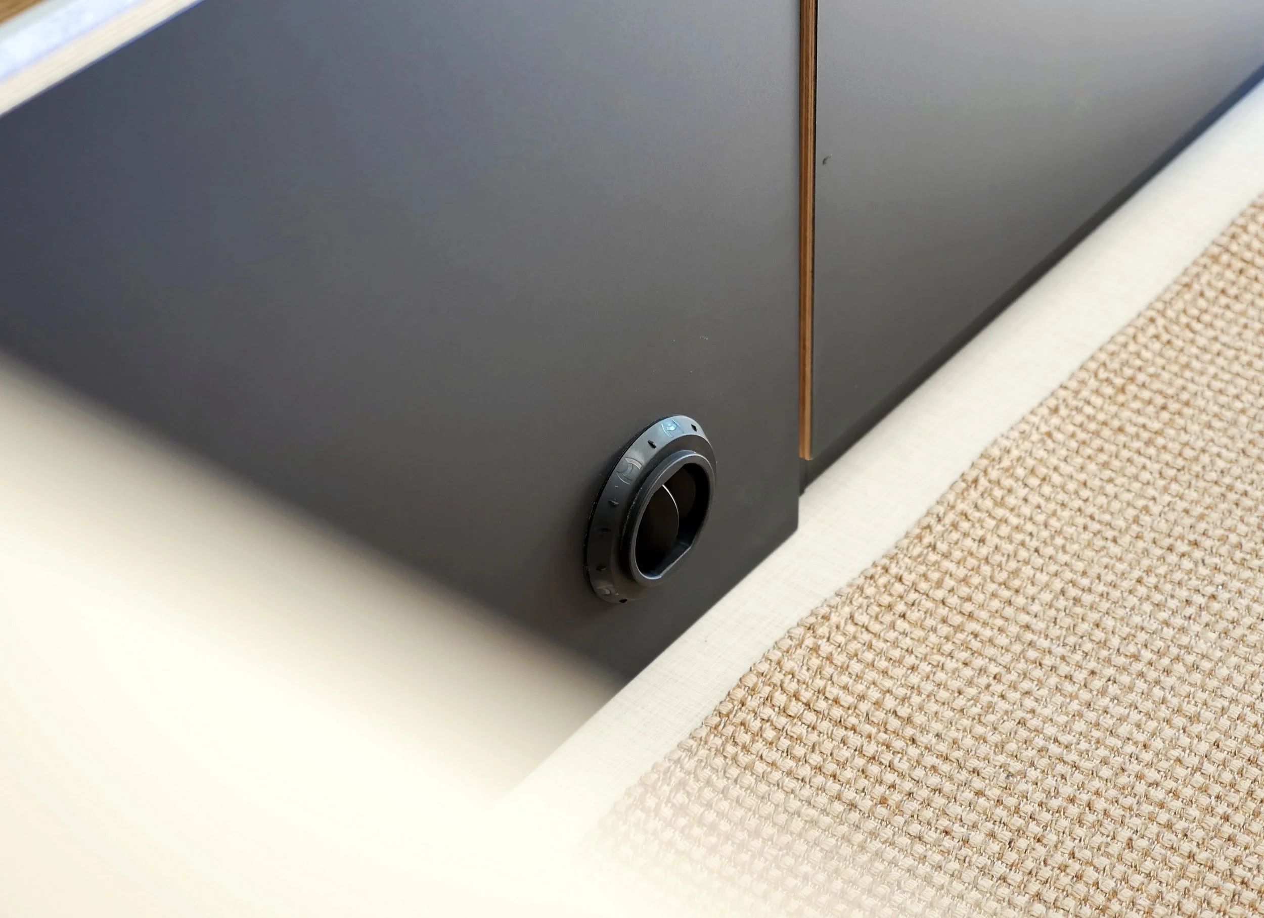 Custom van conversion hardware detail in 'Bjorn' — round black recessed heater vent grille set into a dark charcoal cabinet panel beside the natural linen bench cushion, birch plywood edge detail and soft raking light.
