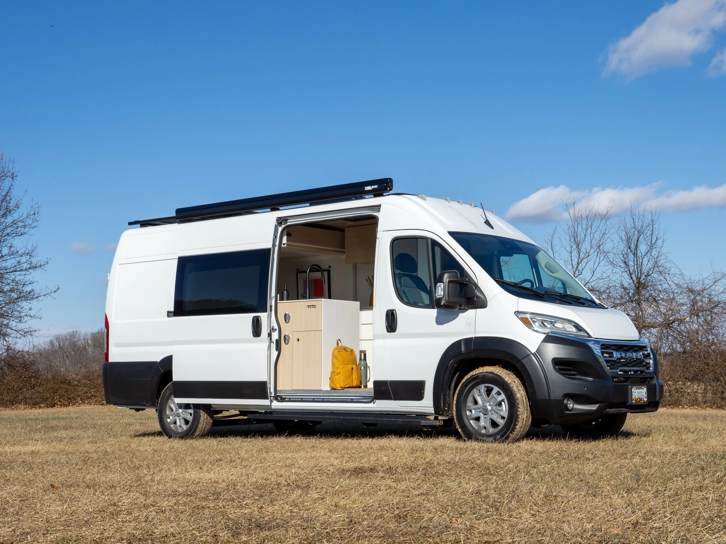 White Ram ProMaster van conversion 'Frida' parked in a winter field with the sliding door open — a custom campervan build by Roamer Vans of Maryland; birch plywood cabinetry and yellow backpack visible inside, black roof rack with solar above, dry la