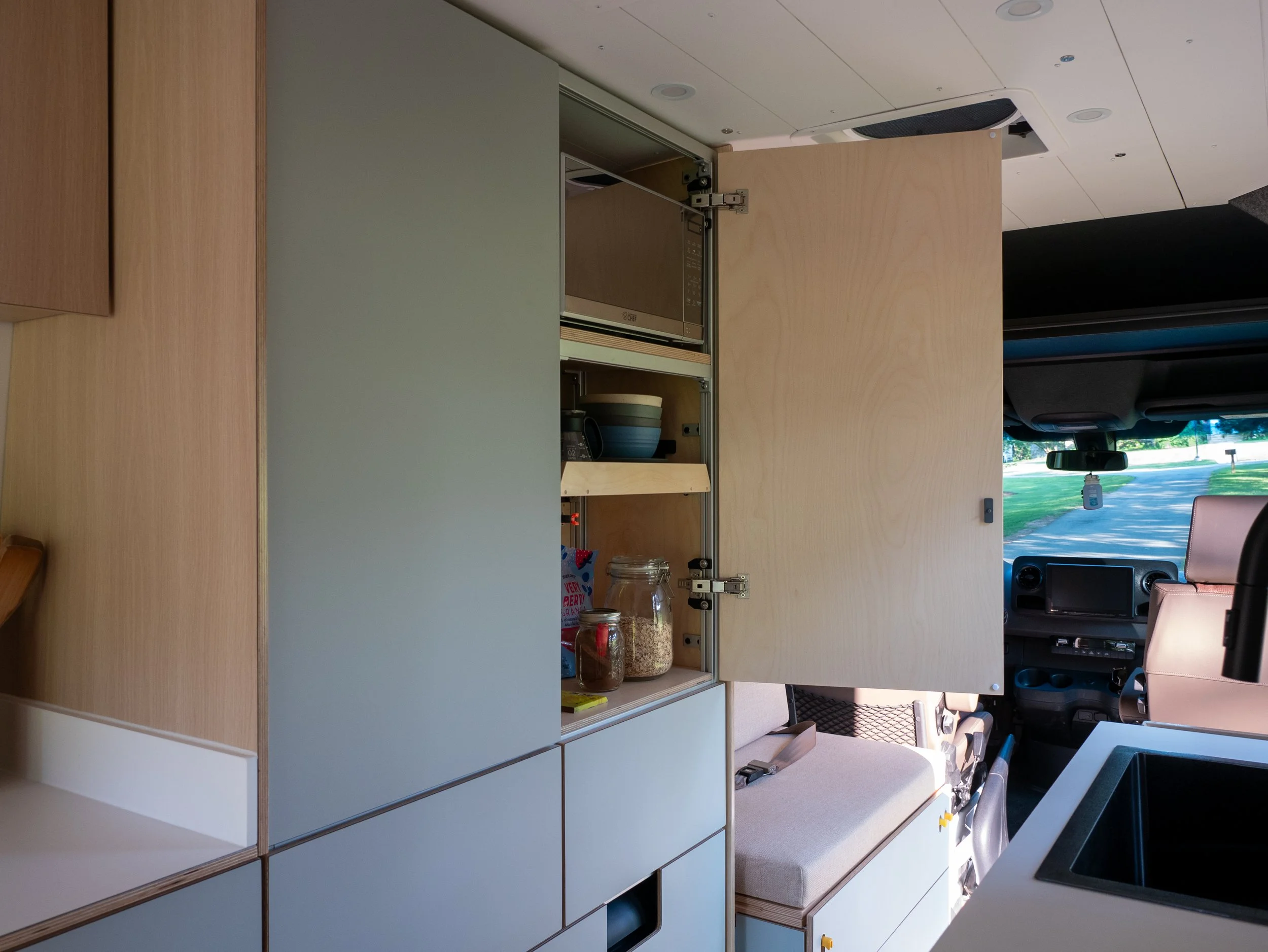 Custom van conversion pantry in 'Aina' — matte sage-green flat-front cabinet doors swung wide to reveal birch plywood shelving with a microwave, stacked bowls, and mason jar pantry storage; bench seat and Sprinter cab visible beyond.