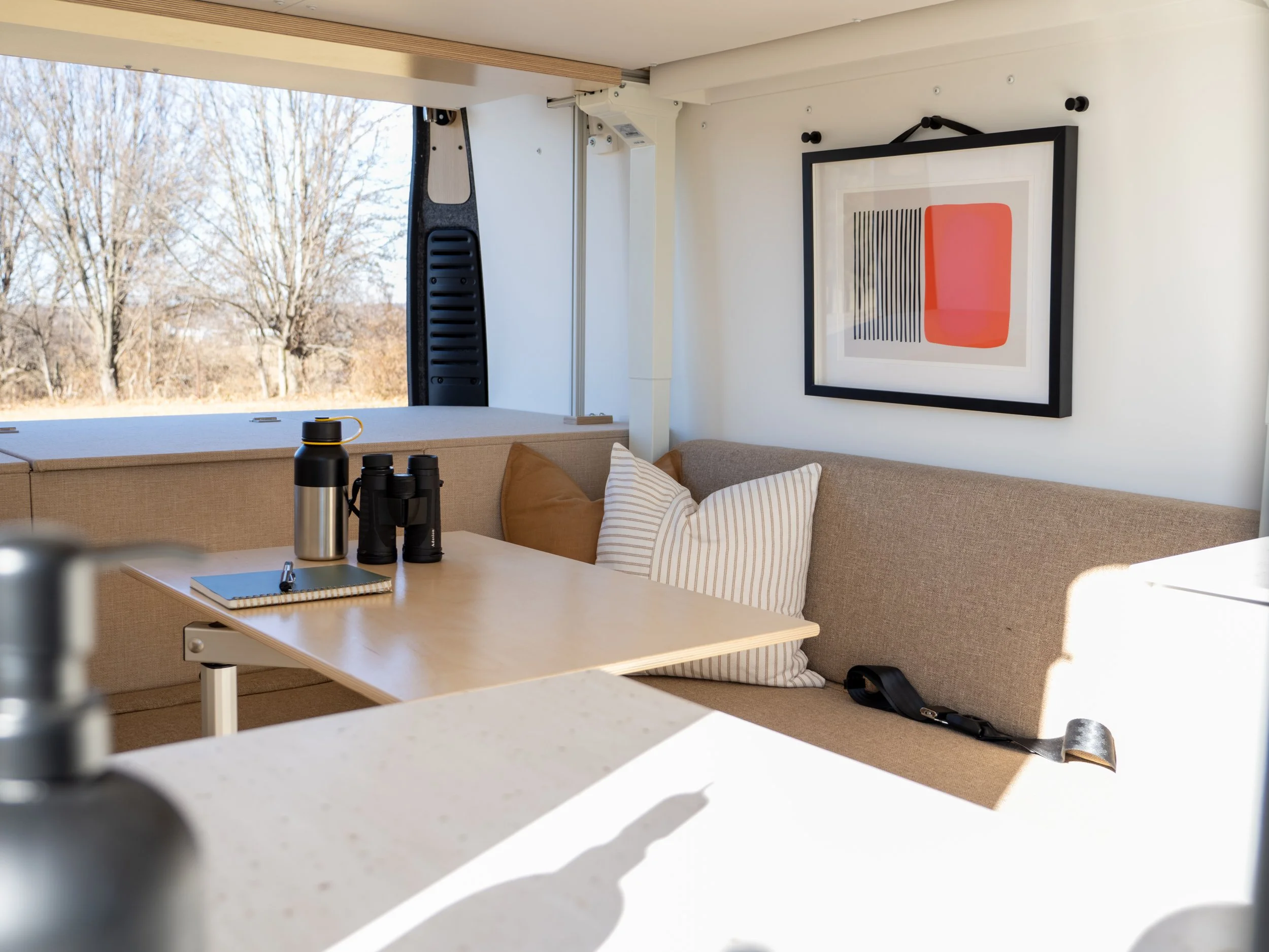 Van conversion lounge in 'Frida' — dinette bench seat with beige upholstery and striped pillow, Lagun plywood table with Stanley thermos, binoculars and notebook on top; large panoramic rear window with bare winter trees beyond, red and graphic abstr