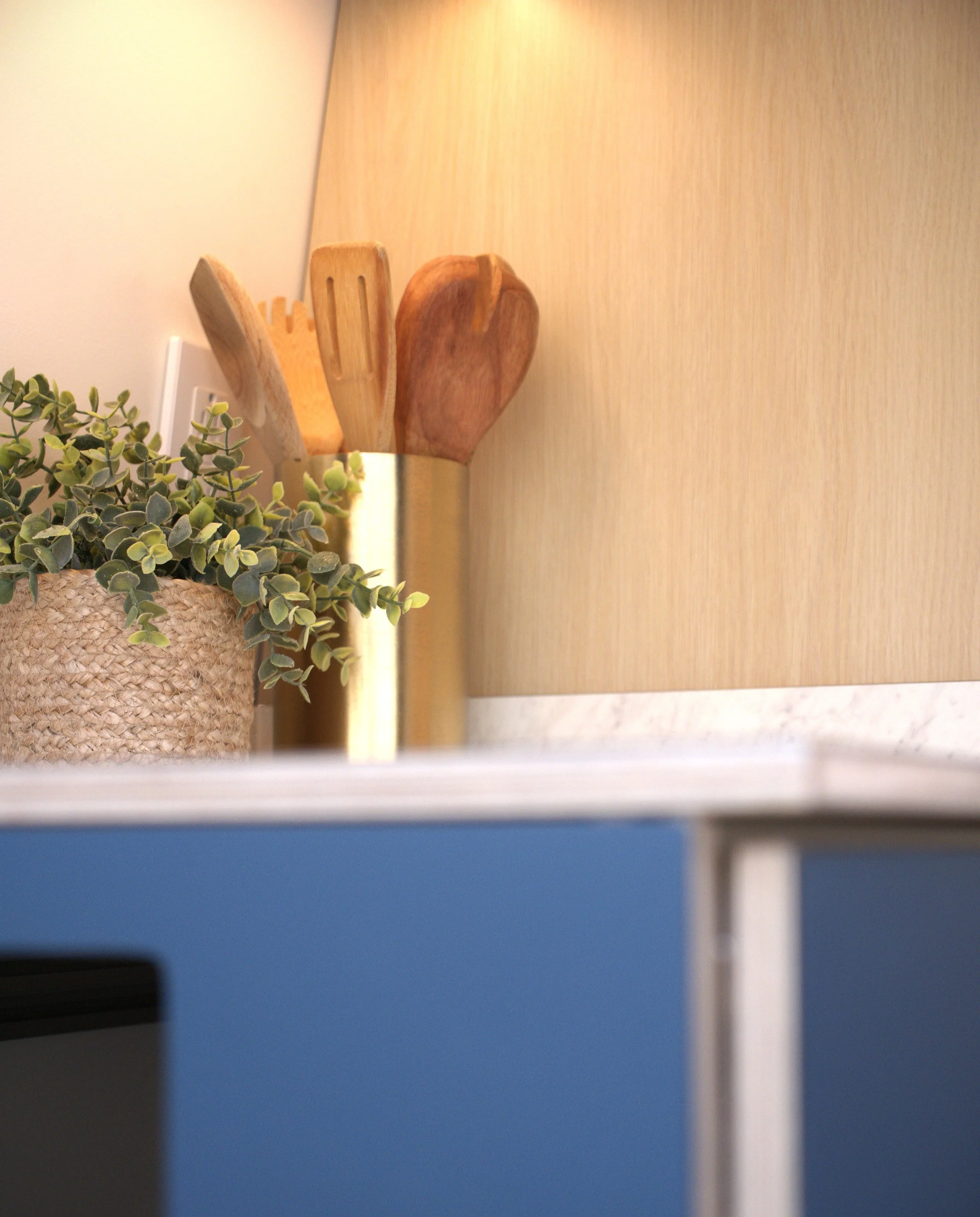 Kitchen counter detail in 'Nordlys' — woven plant pot with trailing greenery and brass utensil holder with wooden spatulas on white countertop, birch plywood cabinet warmly lit from above