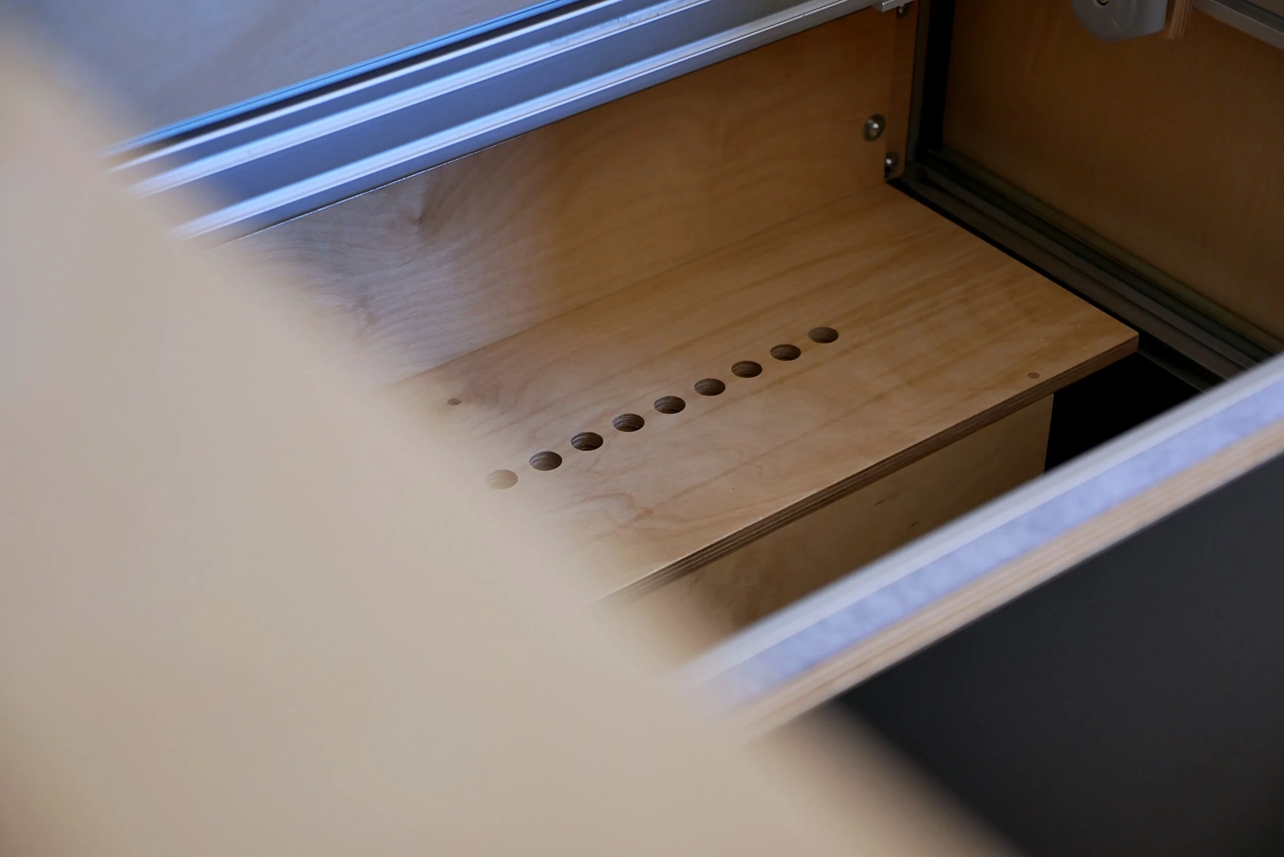 Van conversion heater in 'Bjorn' — aluminum-framed panel open revealing a birch plywood cover for Webasto heater with a precision-drilled row of ventilation holes.