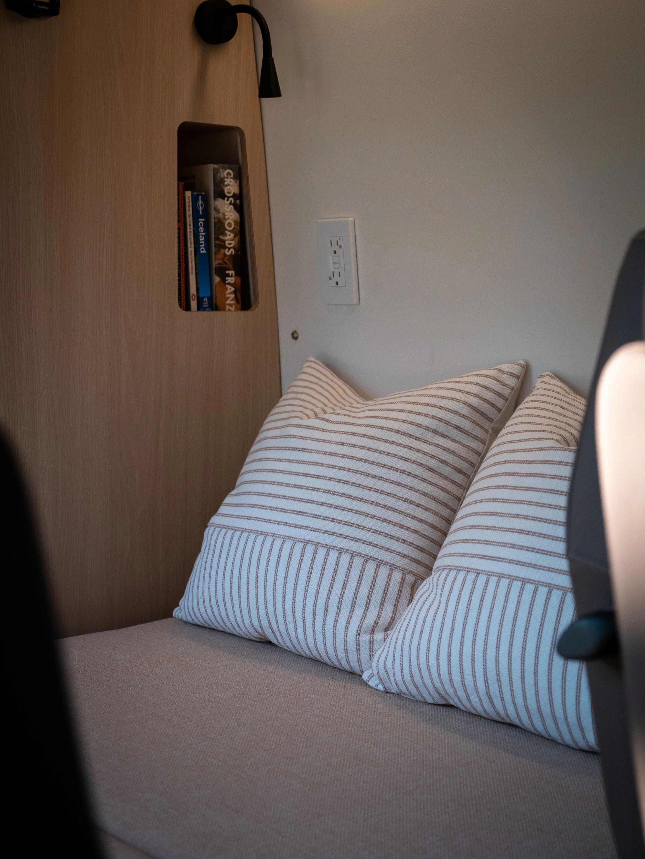 Day-bed and reading nook in 'Aina' van conversion — light linen cushion with red-striped throw pillows, built-in birch wood recessed bookshelf with travel guides, black wall-mounted reading sconce, and a 120V AC outlet in the white wall panel above.