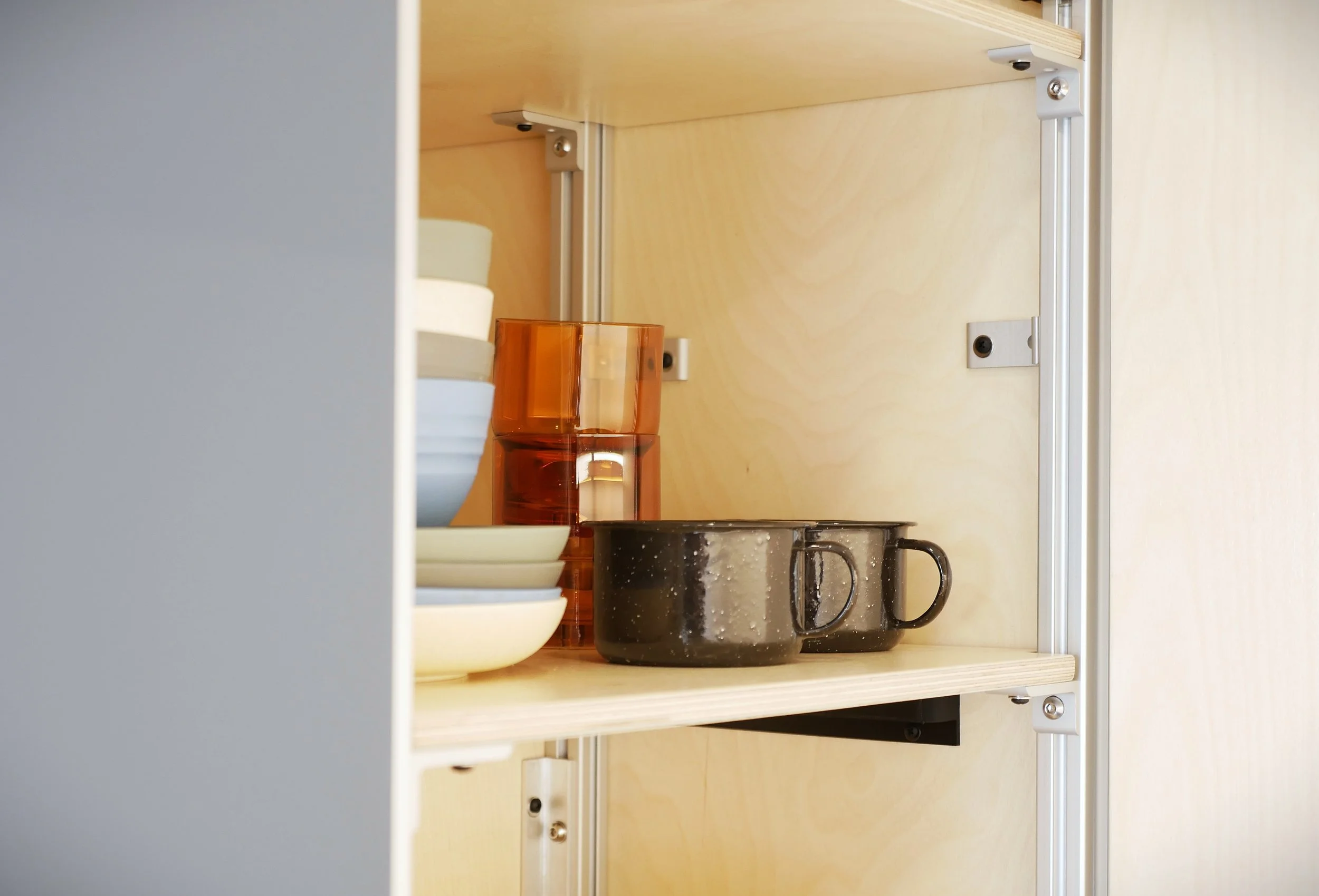 Open kitchen cabinet in 'Nordlys' — plywood-lined shelving with aluminum extrusion framing stocked with speckled ceramic mugs, plates, and amber glassware