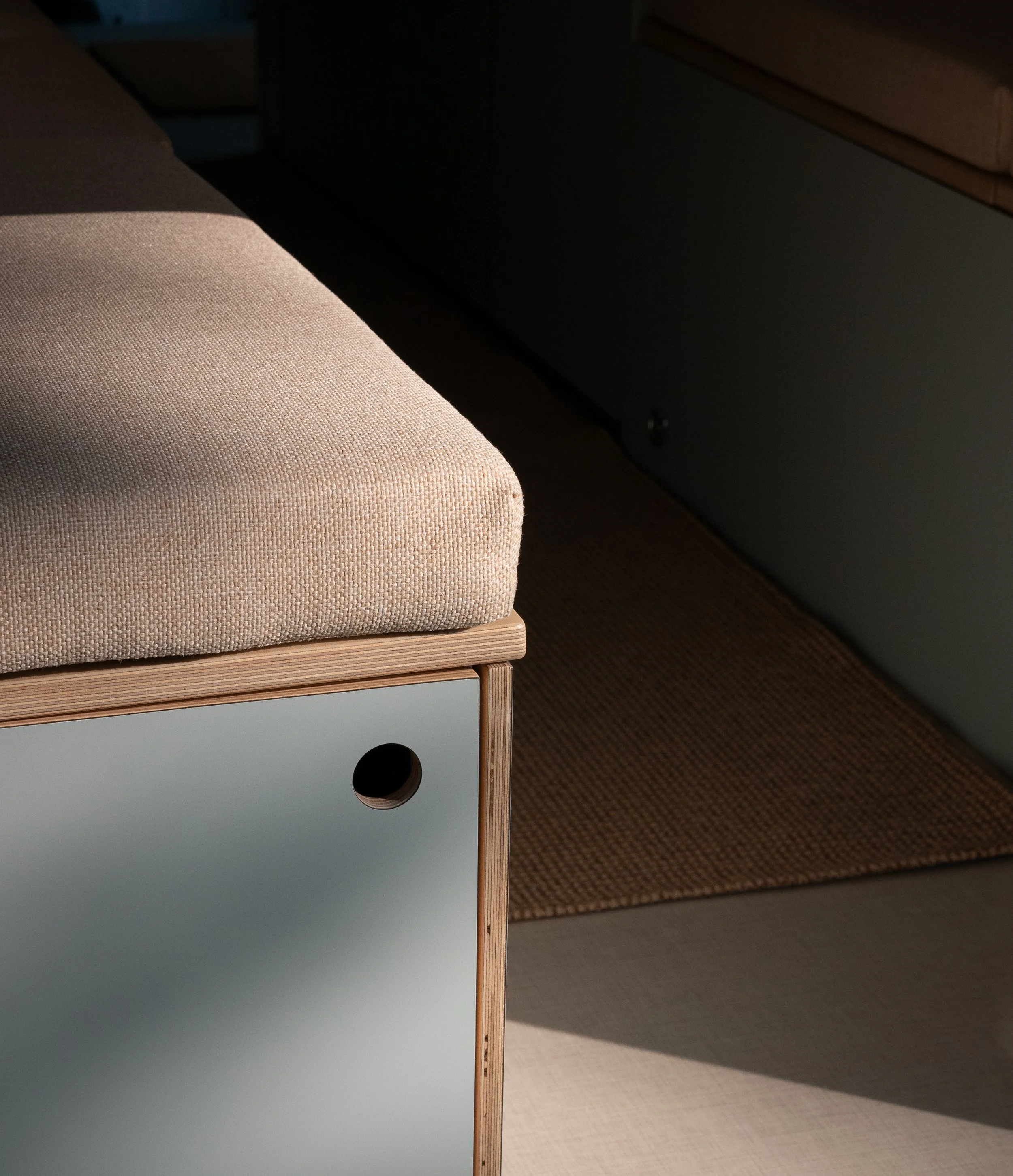 Custom van conversion seating detail in 'Aina' — natural oat color bench cushion on a birch plywood frame with a sage-green flat-front panel featuring a routed circular finger-pull below, light beige vinyl flooring and woven sisal runner at the base,