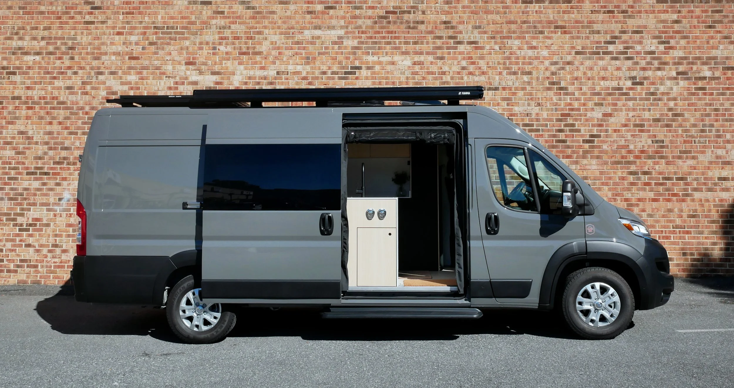 Gray Ram ProMaster high-roof van conversion 'Bjorn' parked with sliding door open at the Roamer Vans Maryland workshop — light wood interior sink cabinet module and light wood floors visible inside, black roof rack with solar panels above.
