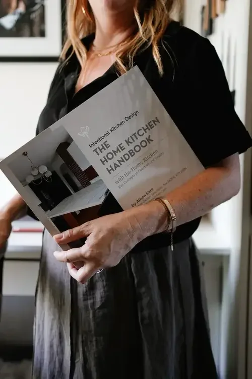 Woman holding a book titled 'The Home Kitchen Handbook' in a room with white walls and shelves.