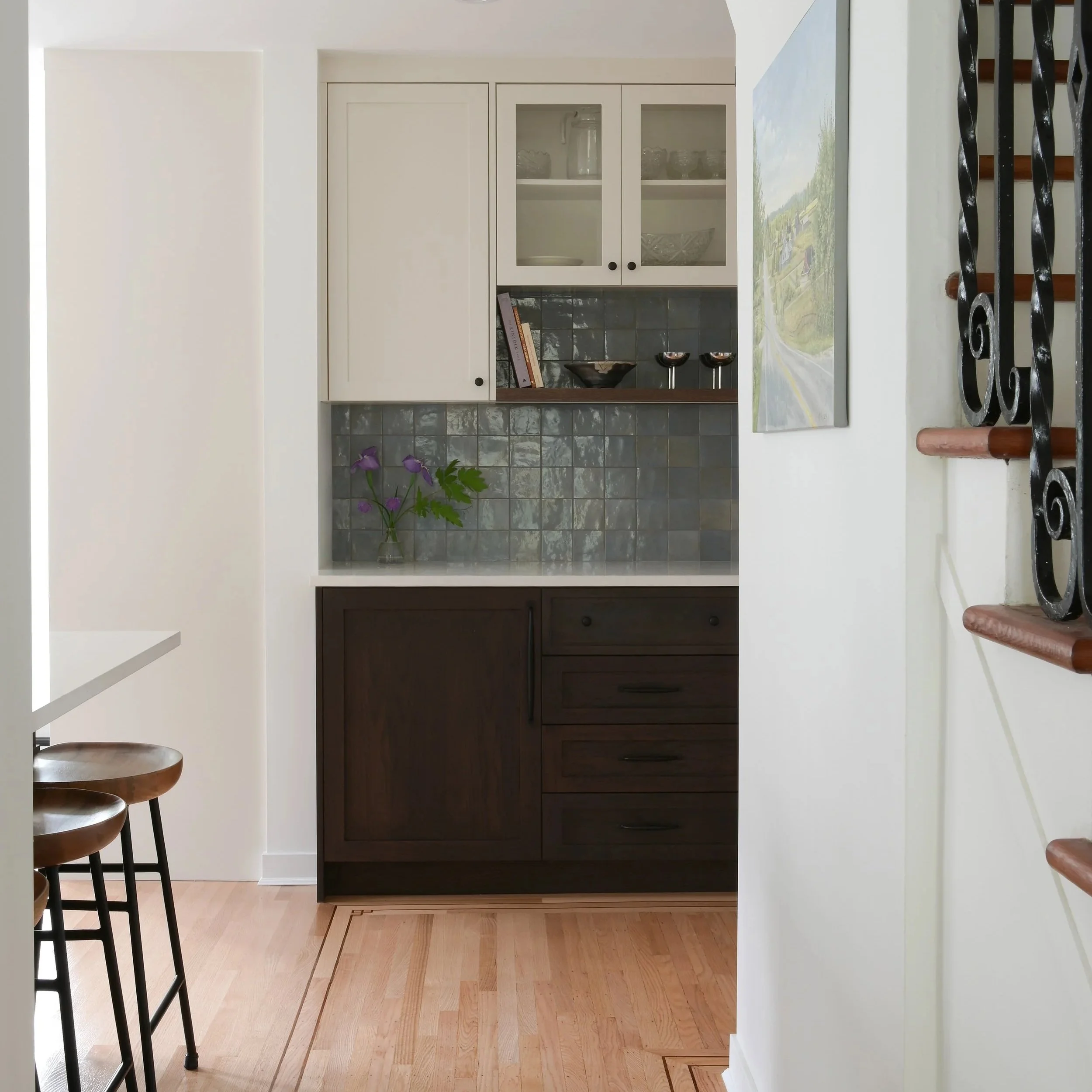 A kitchen area with a white cabinet with glass doors, a black backsplash, and a wooden countertop. There is a purple flower in a glass vase and a small book on the counter. A part of a staircase with wooden steps and black iron railing is visible on the right. The floor is light-colored wood, and a painting or photograph of a rural road with green trees and fields hangs on the wall.