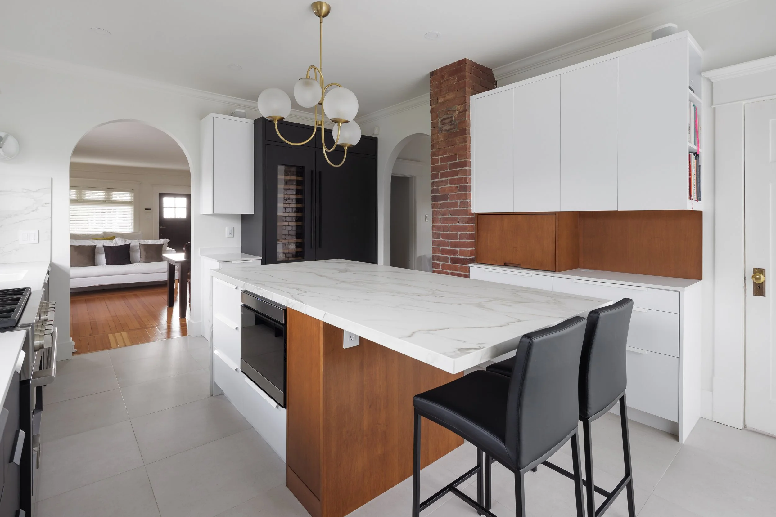 Modern kitchen with white cabinets, a marble island, black bar stools, and a brick column. A chandelier hangs from the ceiling, and a doorway leads to a living room with a couch.