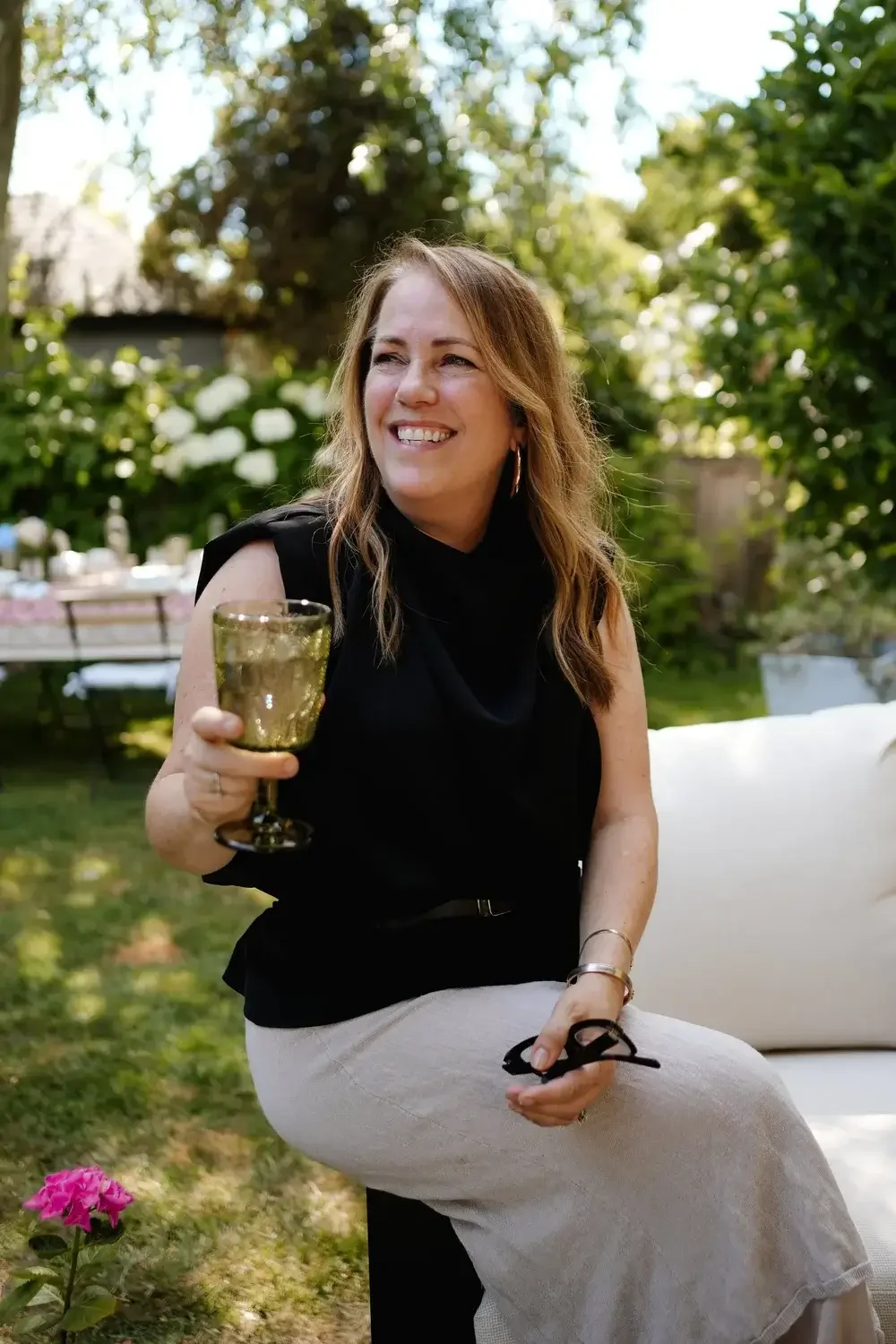 A woman sitting outdoors in a garden during daytime, holding a glass of wine, smiling, dressed in a black top and light-colored skirt.