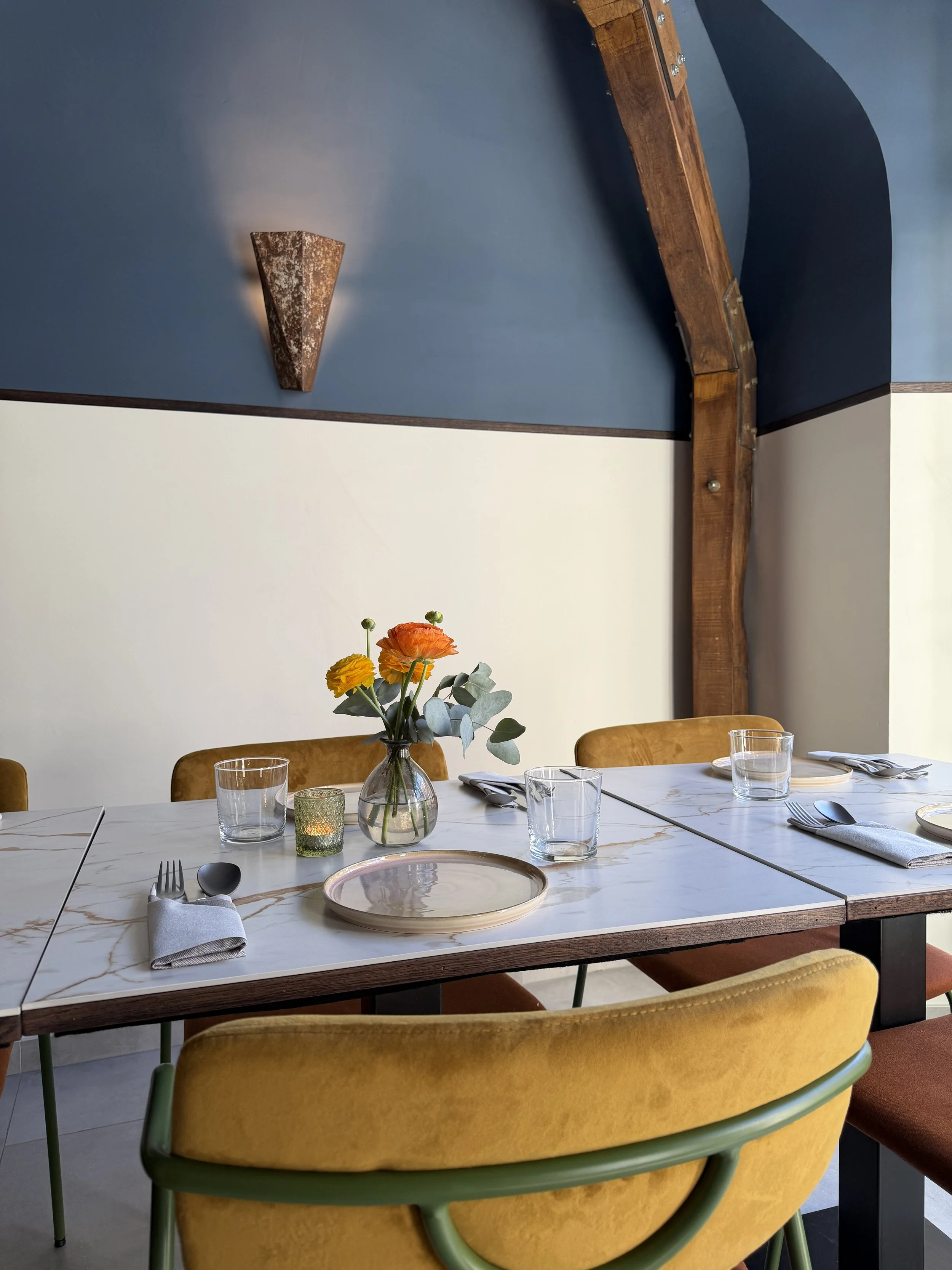 A dining table set for a meal with plates, glasses, utensils, a candle holder, and a flower vase with orange and yellow flowers. Surrounded by mustard-colored chairs with green legs. The wall behind is painted blue with a wooden beam and a wall sconce.