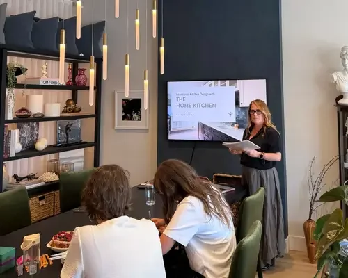 A woman presenting to a small group in a modern meeting room with a TV screen, shelves with decor, and plants.