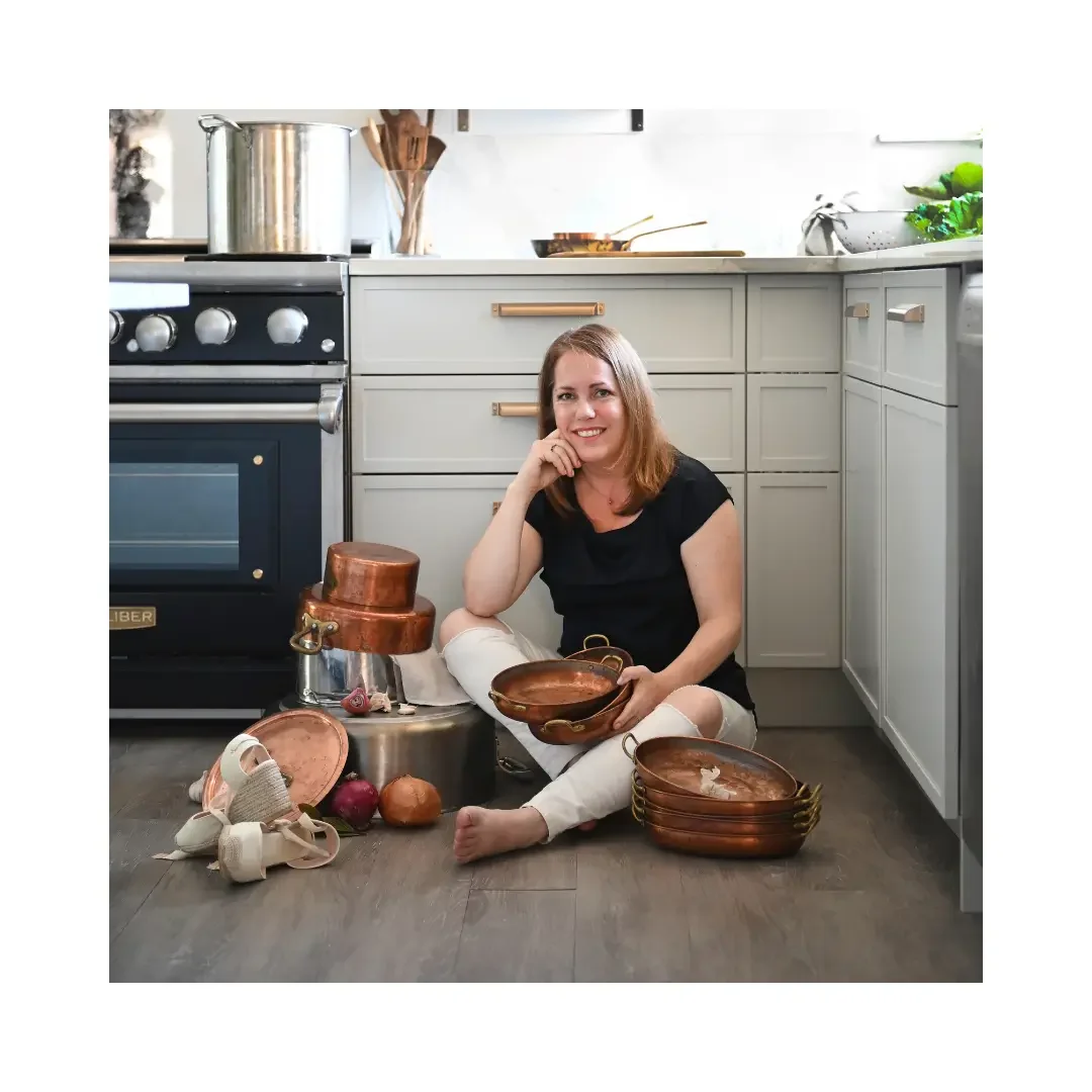 A woman sits on the floor surrounded by various pots and pans, appearing engaged in cooking or organizing kitchenware.