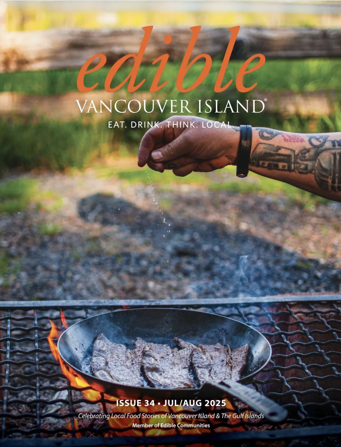 A hand with tattoos on the arm sprinkling salt over a sizzling piece of meat in a frying pan on an outdoor grill. The background is blurred with a wooden fence and greenery.