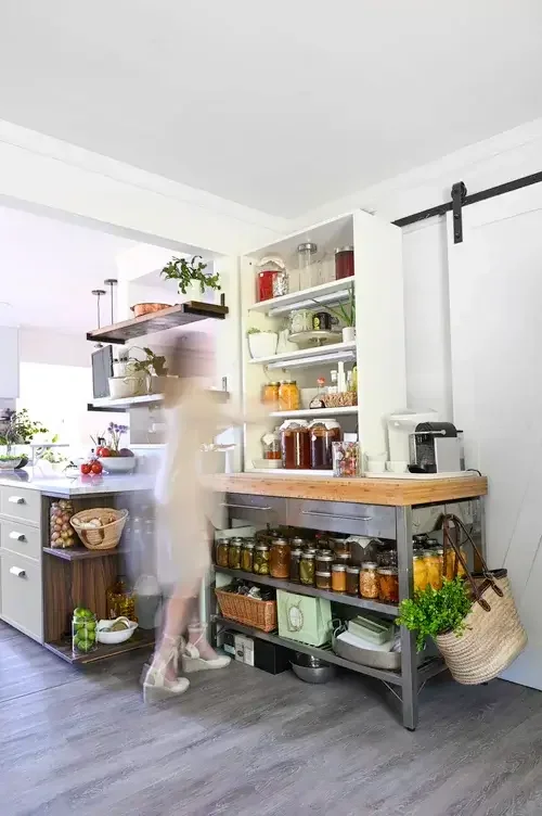 A woman walks through a kitchen filled with various food items, showcasing a vibrant and bustling cooking environment.