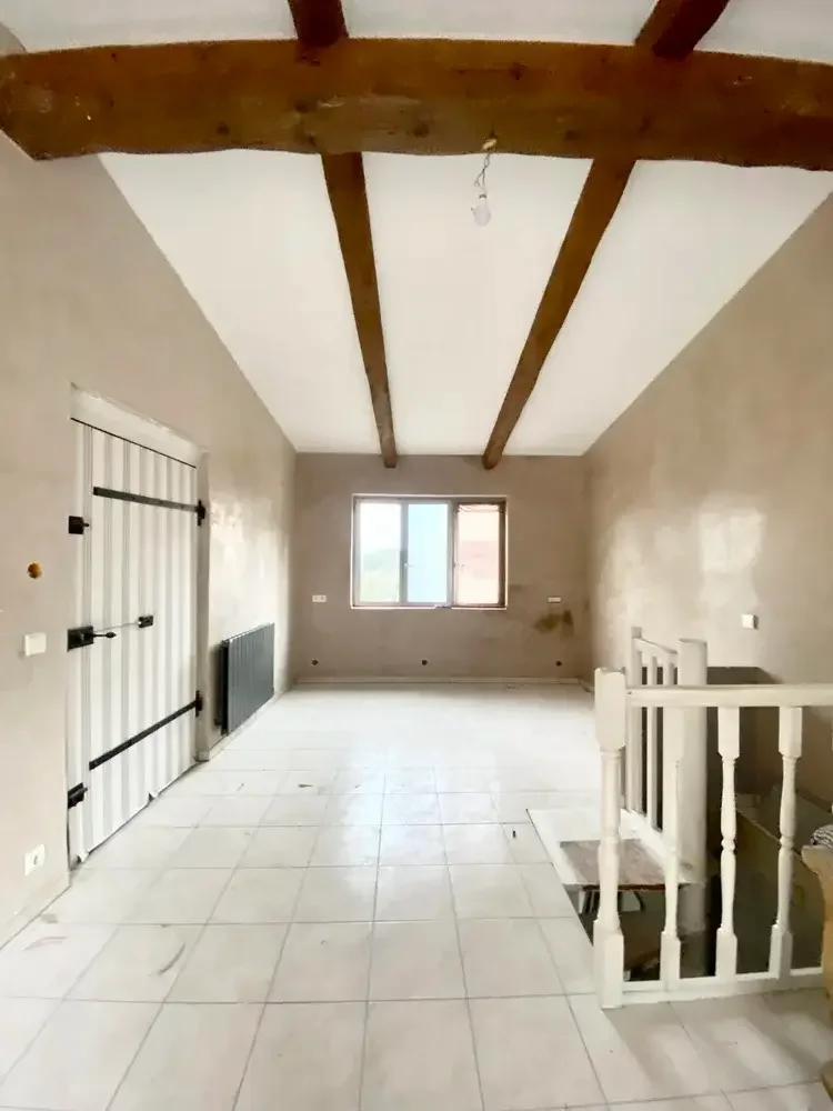 An empty room with beige painted walls, white tiled floor, exposed wooden ceiling beams, a window on the far wall, a sliding barn door on the left, a radiator, and a staircase with a white railing leading downstairs.