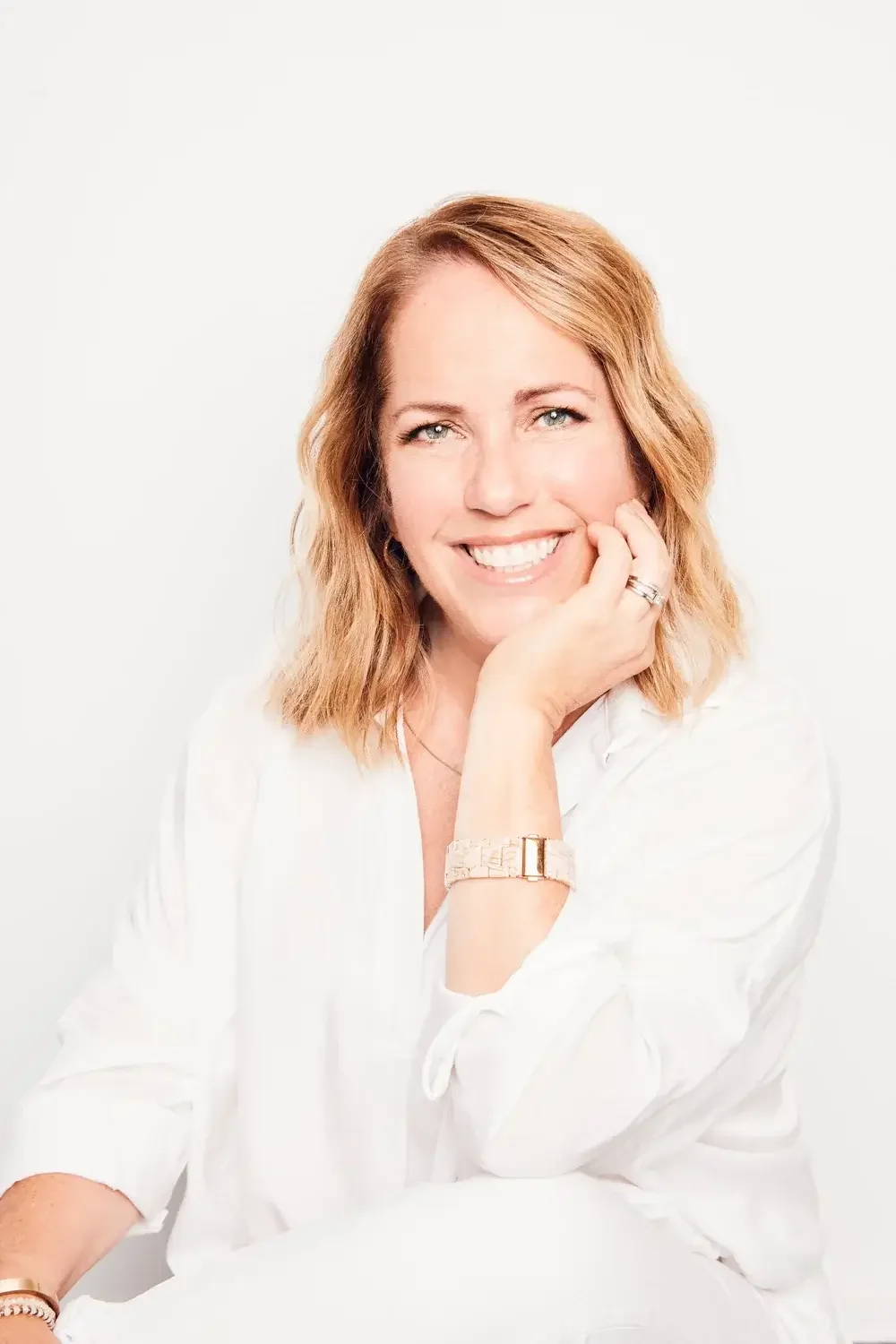 A woman with blonde, shoulder-length hair, smiling, resting her chin on her hand, wearing a white shirt and jewelry, against a plain white background.