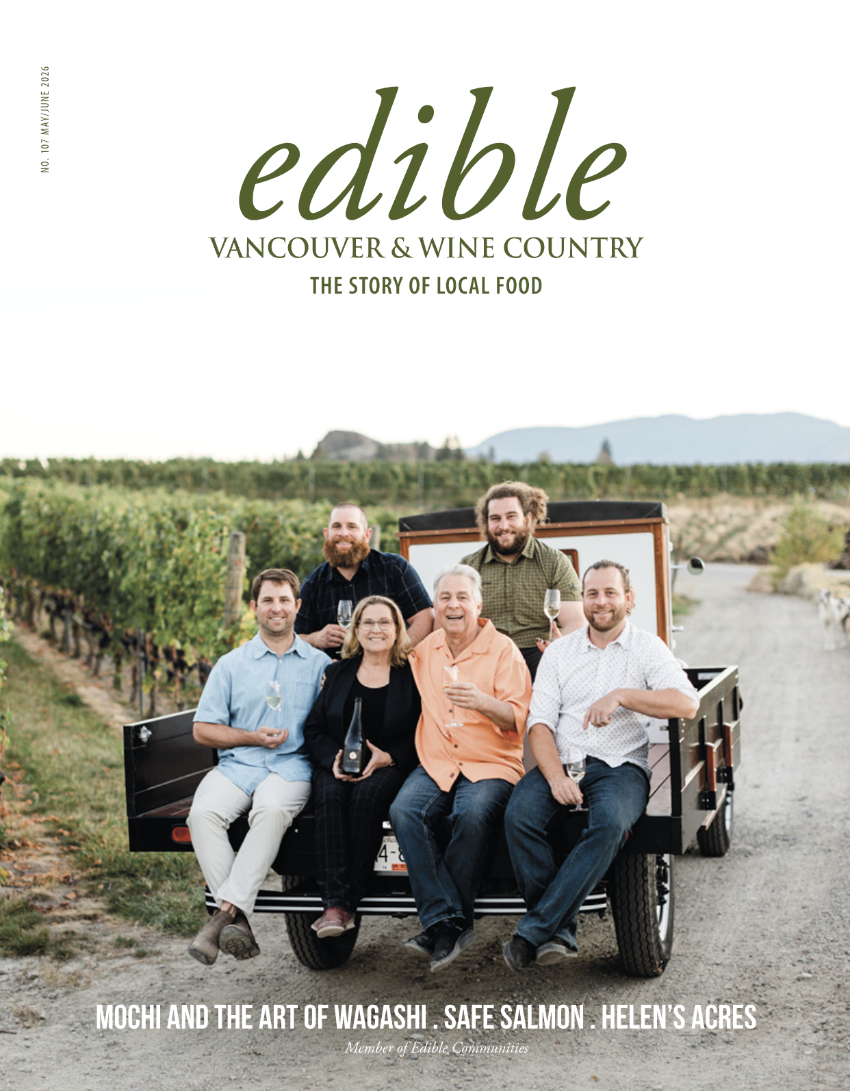 A group of six people sitting and standing on a small vehicle in a vineyard, holding drinks and smiling, with a mountain in the background.