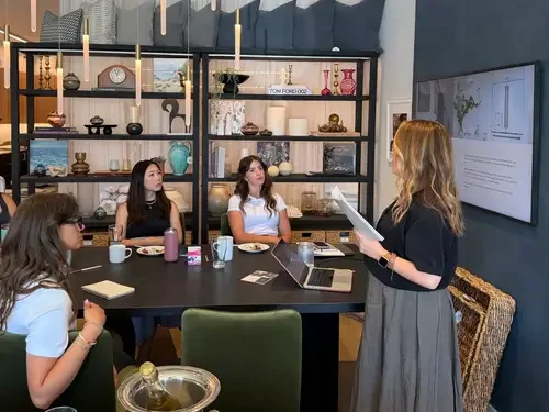 A woman standing and presenting to four young women seated at a meeting table, with a large screen displaying a presentation on the wall and a bookshelf in the background.