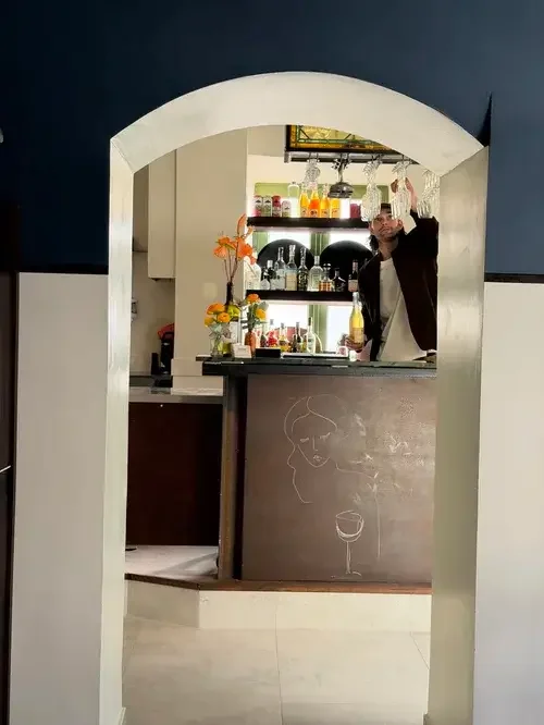 A man stands confidently in front of a bar, showcasing a casual yet stylish demeanor.
