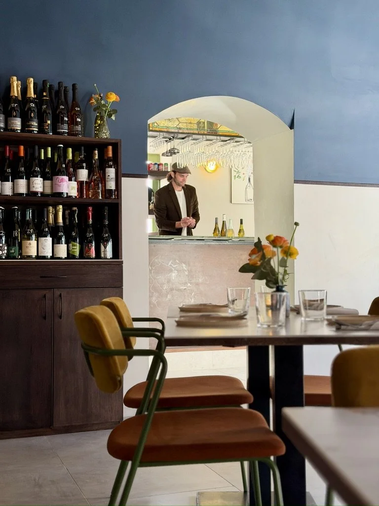 A man in a brown blazer and hat preparing drinks behind the bar at a restaurant with a blue and white interior, a wooden shelf with wine bottles, and a dining table with chairs and a flower vase.