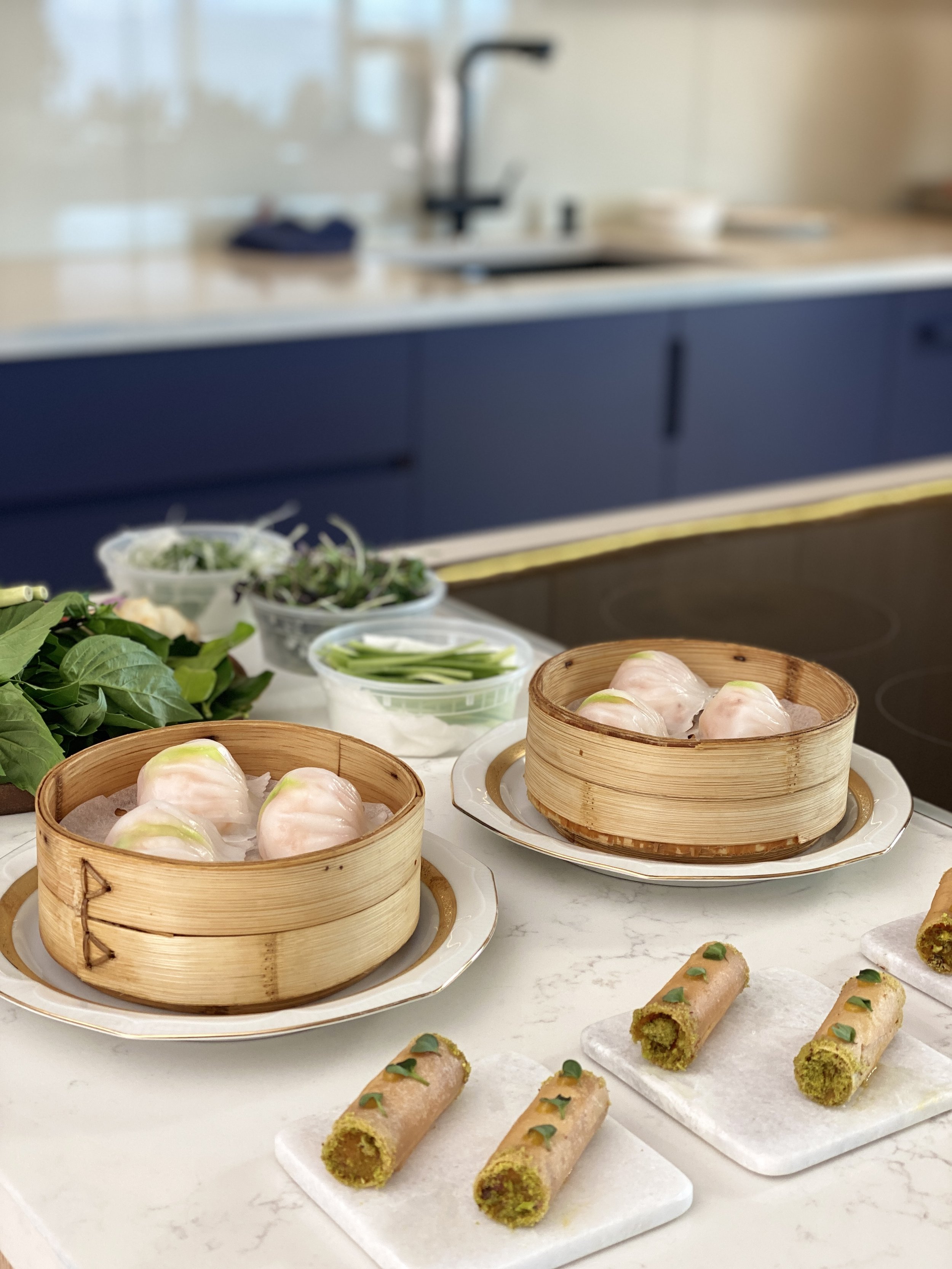 Table with bamboo steamers containing dumplings, alongside plates of green onion and other garnishes, with some rolled appetizers on a white surface in a bright kitchen.
