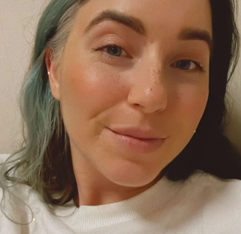 A close-up photo of a woman with mixed green and black hair, light skin with freckles, and a subtle smile, wearing a white shirt and small earrings.