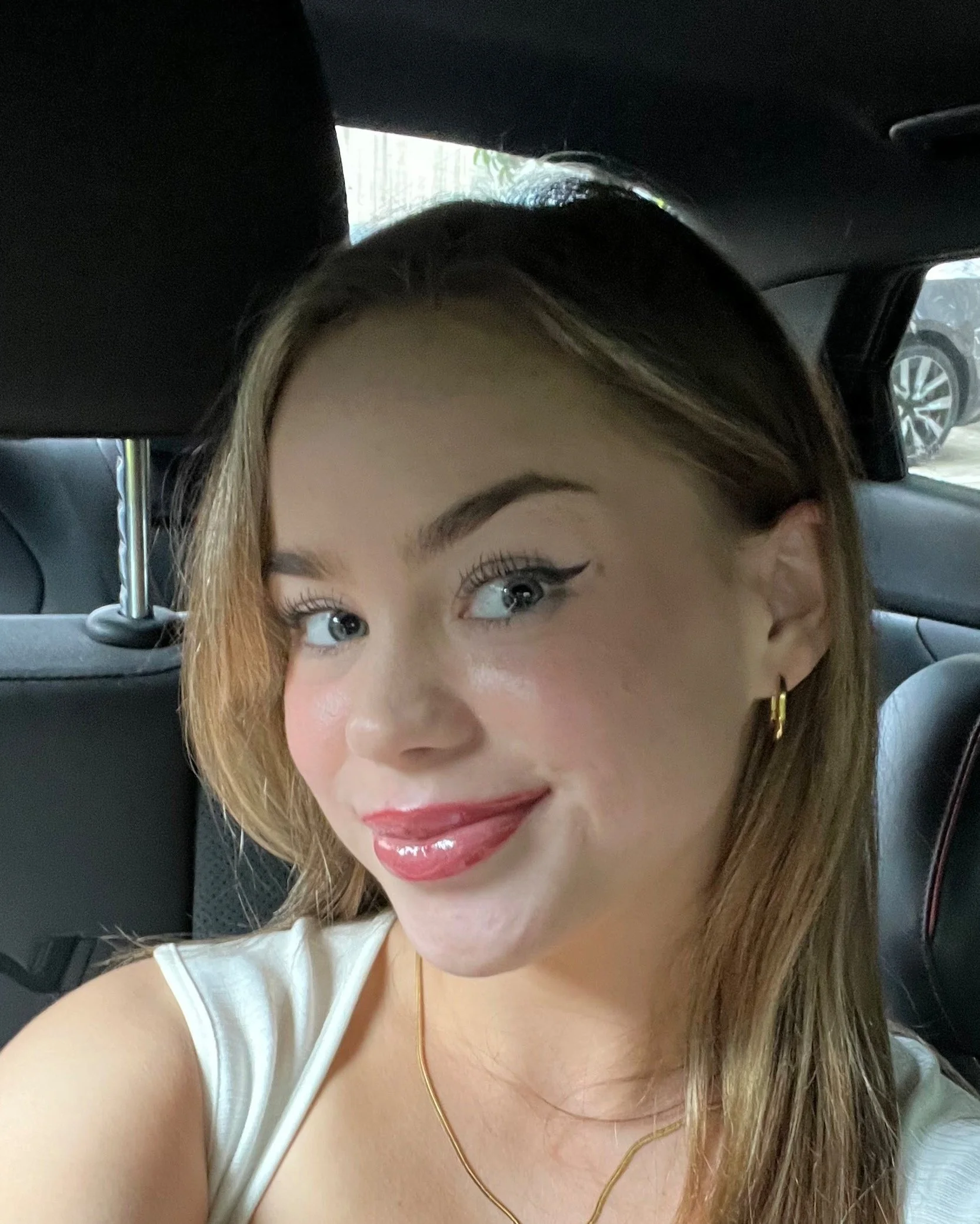 Close-up of a young woman with brown hair, blue eyes, and makeup, smiling in a car.