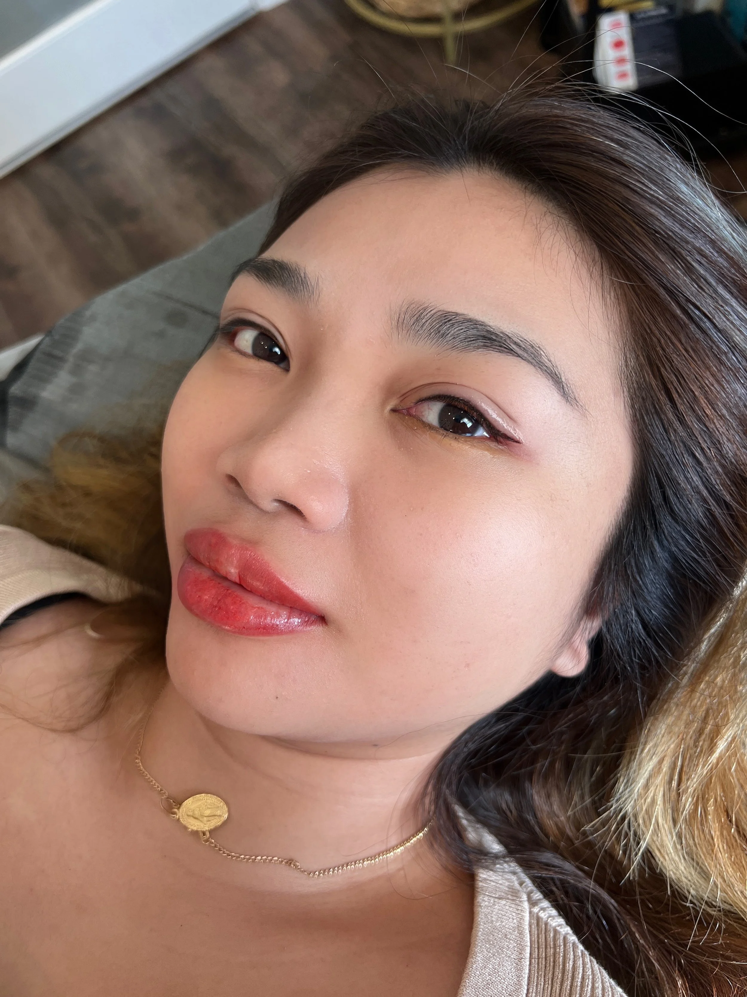 Close-up of a woman with dark hair and makeup, lying on a surface with a gold necklace and a beige top, with a wooden floor and some furniture visible in the background.
