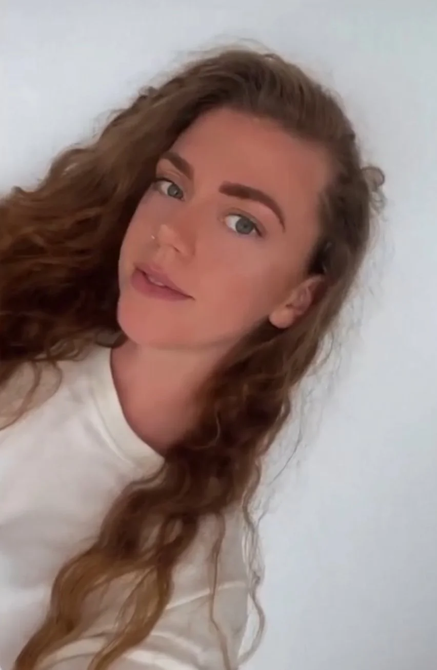 A young woman with long, wavy light brown hair, wearing a white shirt, looking directly at the camera with a slight smile against a plain light background.