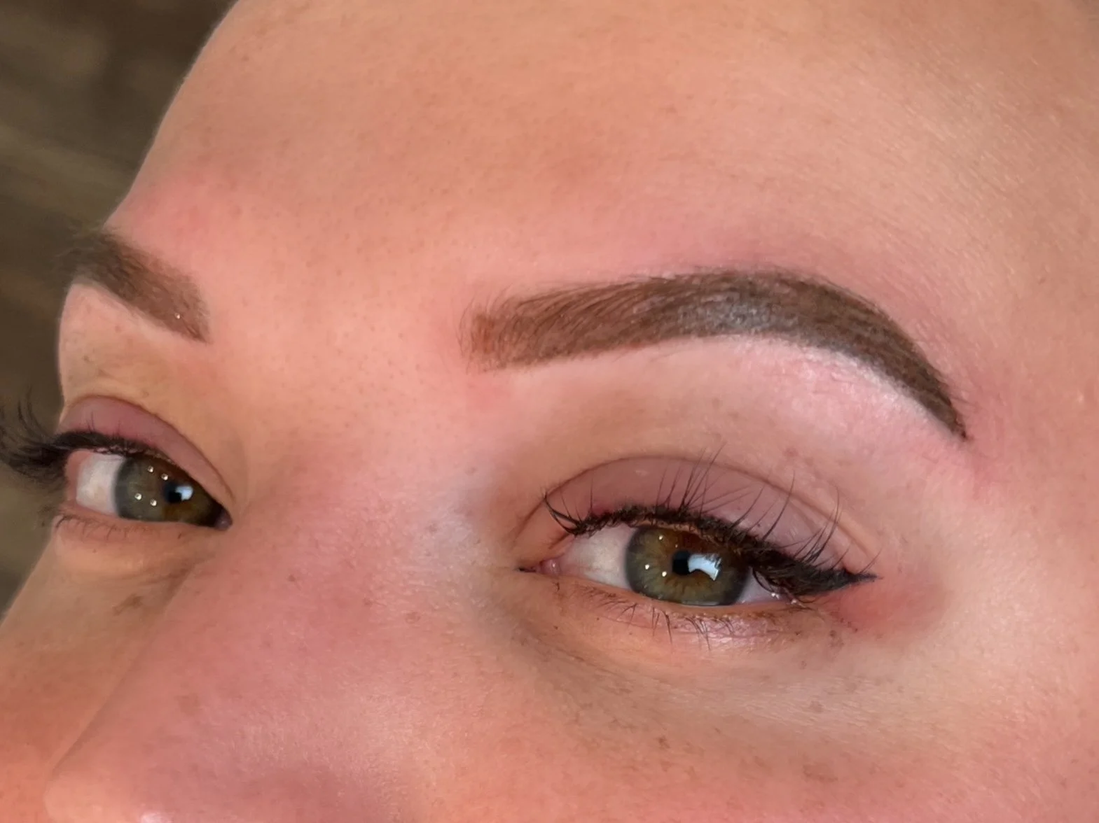 Close-up of a woman's face showing her eyes and eyebrows. She has well-groomed eyebrows, eye makeup with eyeliner and mascara, brown eyes, smooth skin, and some freckles.