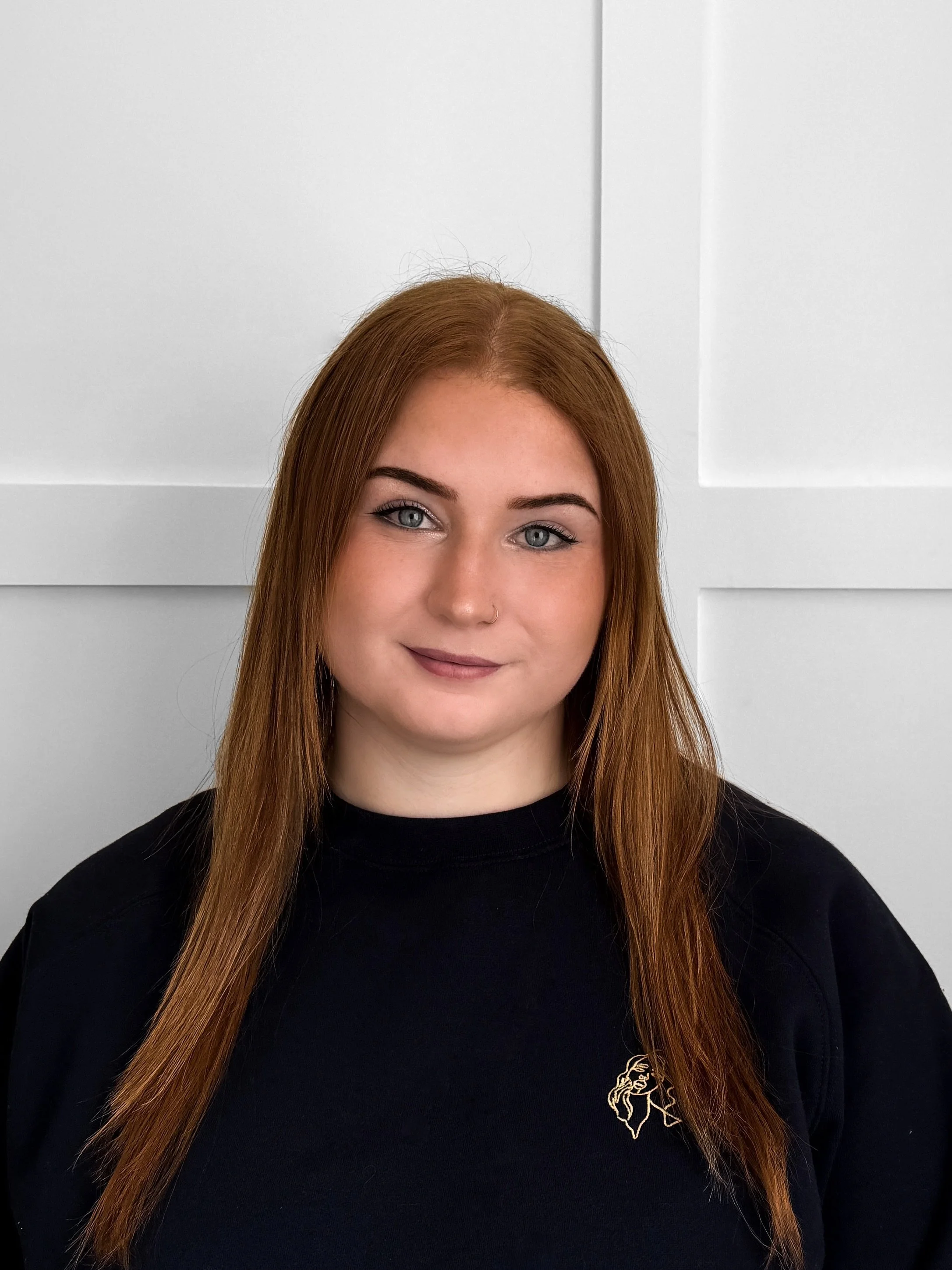 A young woman with long red hair, blue eyes, and a nose piercing smiling at the camera, wearing a black shirt with a small gold logo on the chest, standing in front of a white paneled wall. Allure Ink Staff