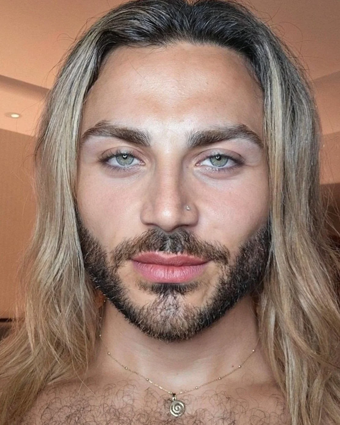 A person with long, blonde hair, light skin, blue eyes, and facial hair, wearing a necklace with a spiral pendant, looking directly at the camera.