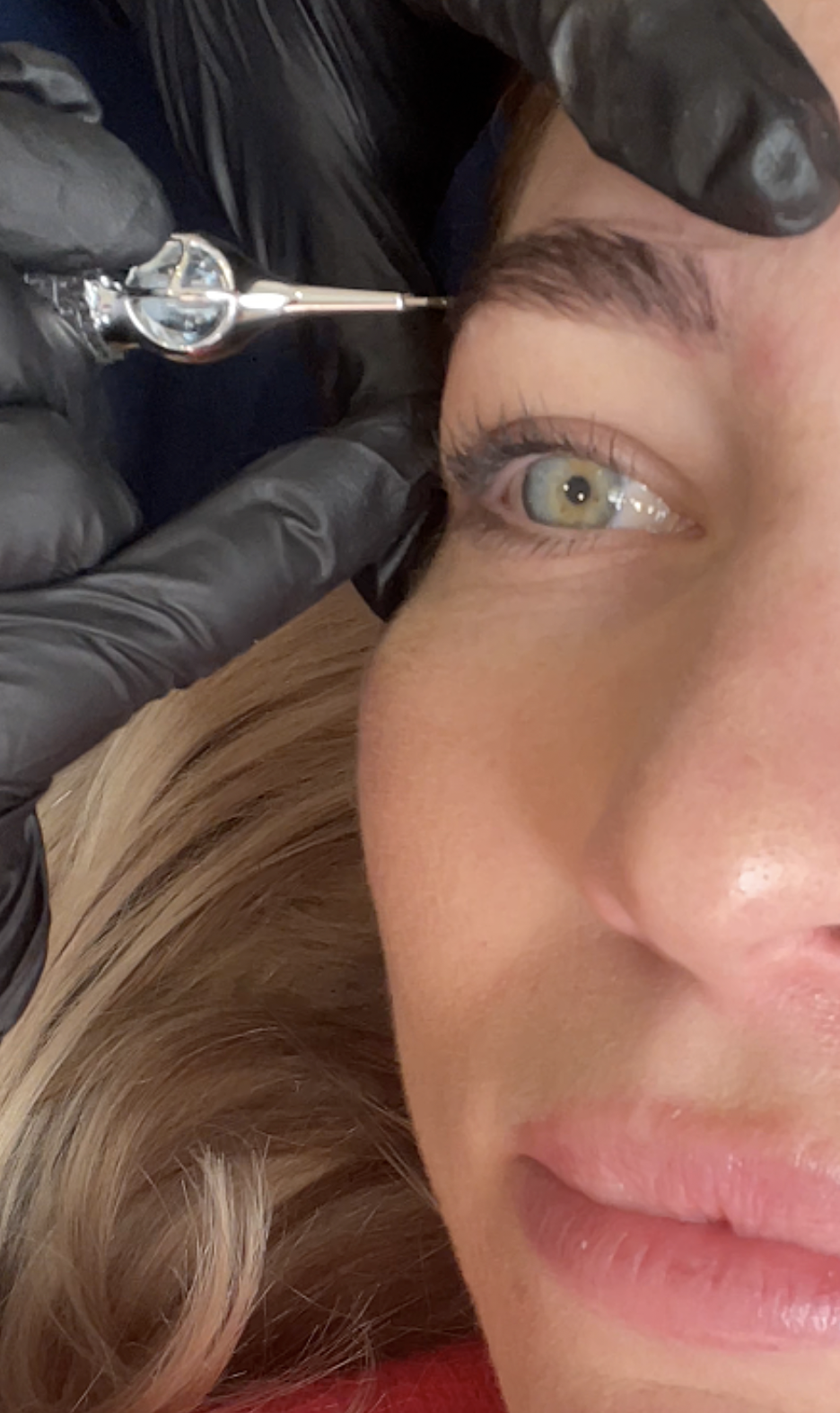 A person with blonde hair getting a tattoo near their eye from a tattoo artist wearing black gloves.