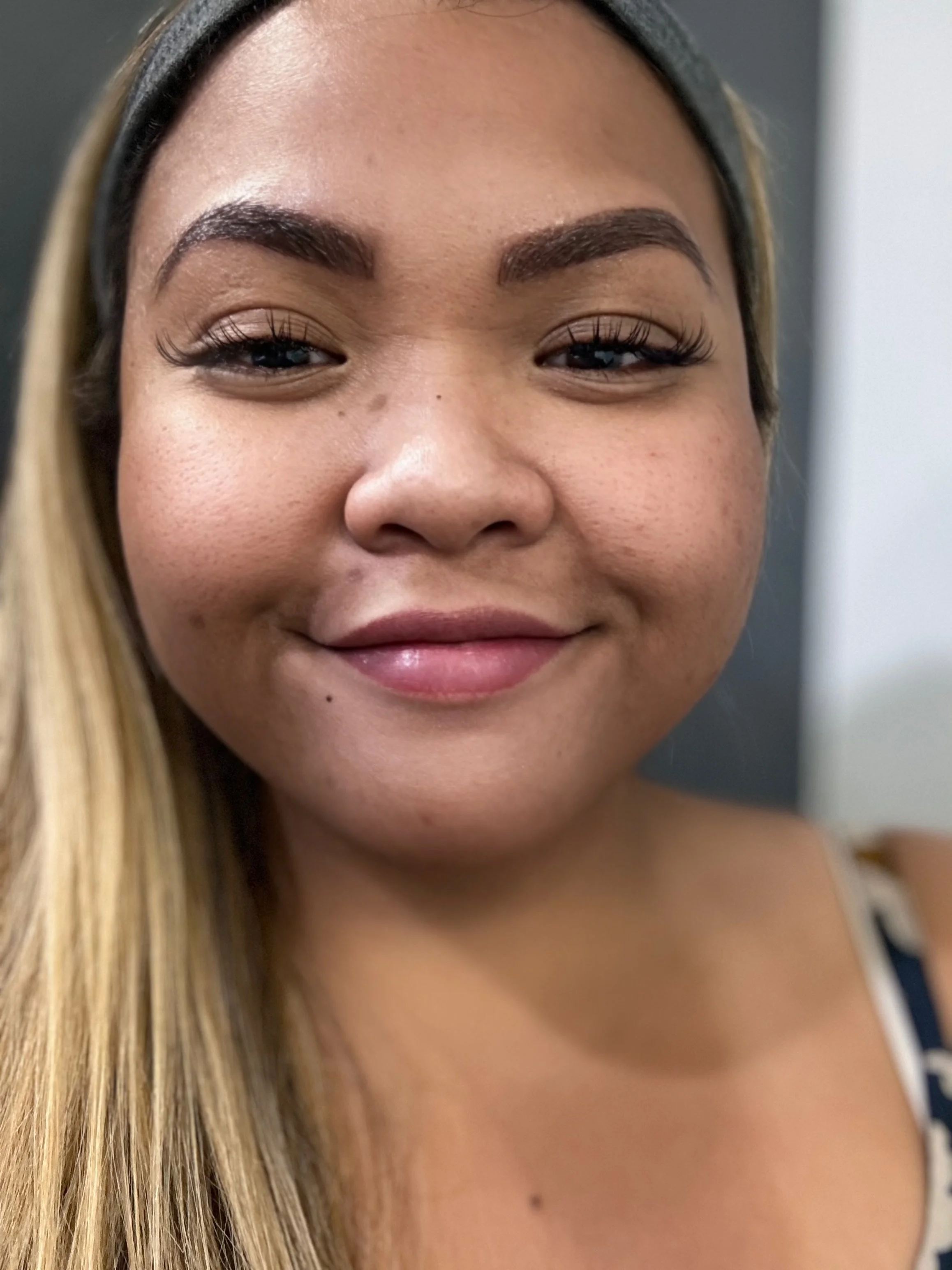 Close-up of a smiling woman with tan skin, long blonde hair, and brown eyebrows, wearing makeup and a black headband, in front of a plain background.