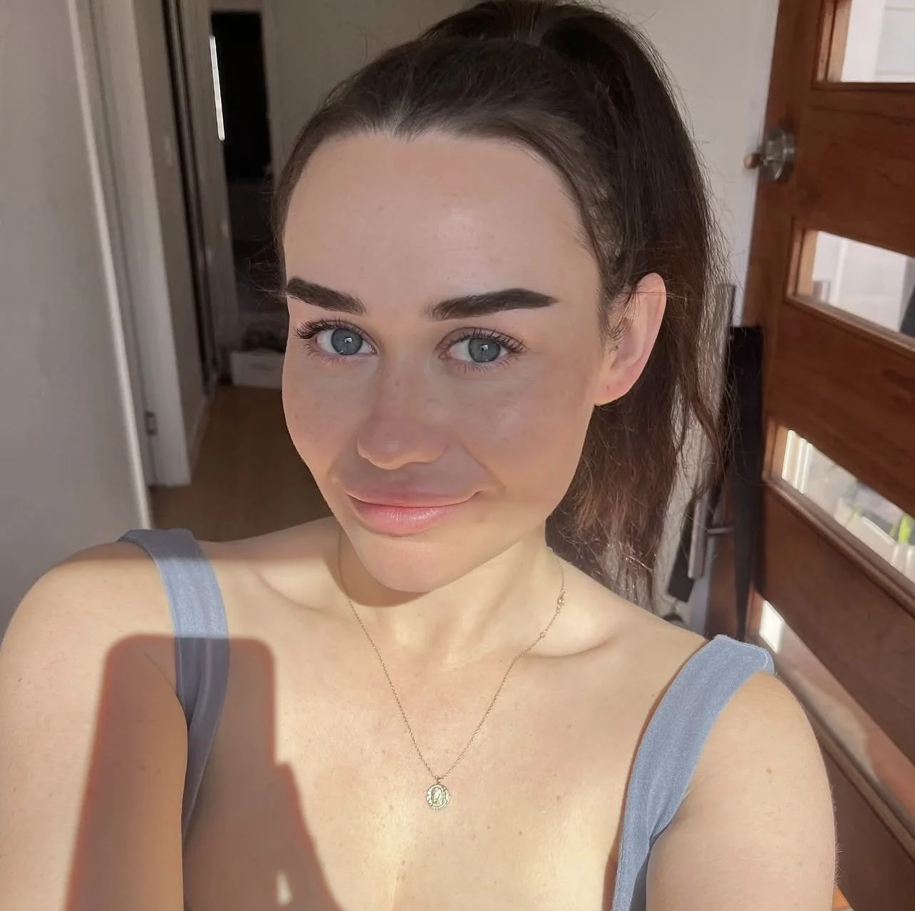 A young woman taking a selfie inside a house, with a bright expression, light blue eyes, and dark brown hair pulled back into a ponytail. She's wearing a gray tank top and a delicate gold necklace with a small pendant.