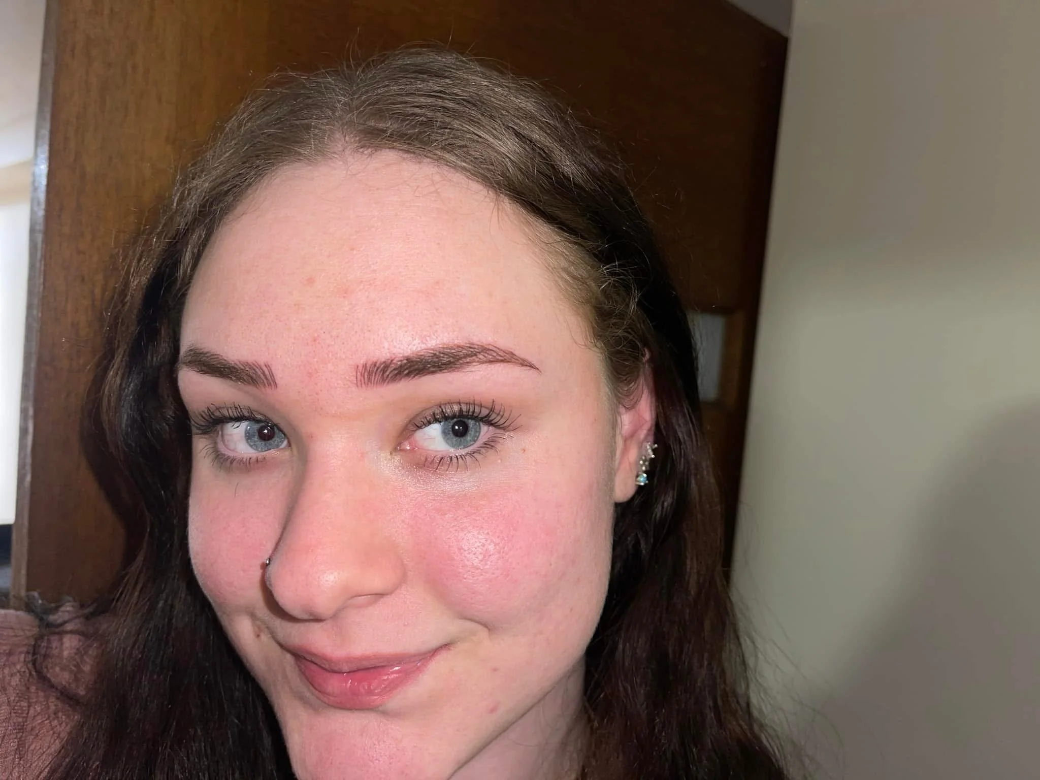 A young woman with long brown hair, blue eyes, and a nose piercing, smiling in a close-up selfie indoors, with a wooden door and wall in the background.