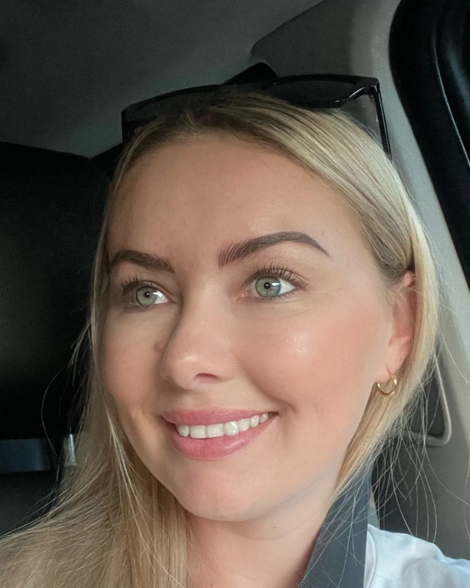 Close-up of a woman with blonde hair, blue eyes, wearing makeup and gold hoop earrings, sitting in a car, smiling and looking to the side.