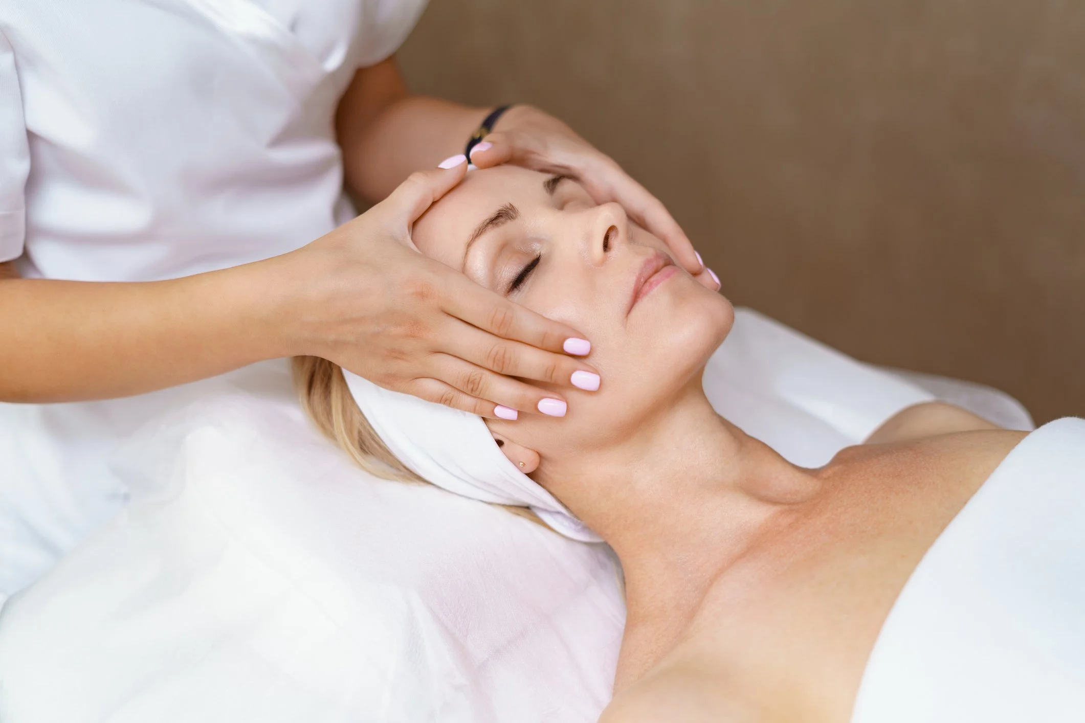 A woman receiving a facial massage while lying down with eyes closed, therapist gently massaging her forehead and cheeks.