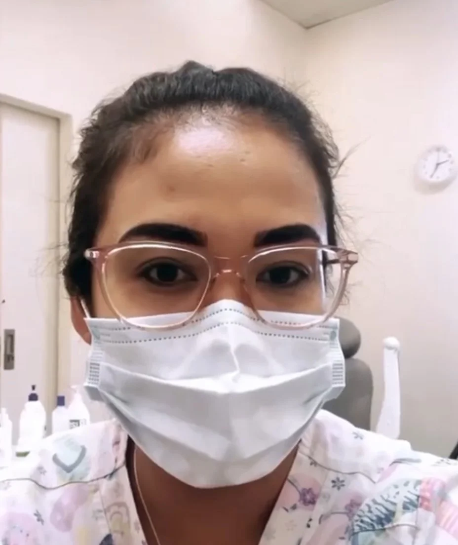 A woman with dark hair tied back, wearing glasses and a white face mask, in a medical setting.