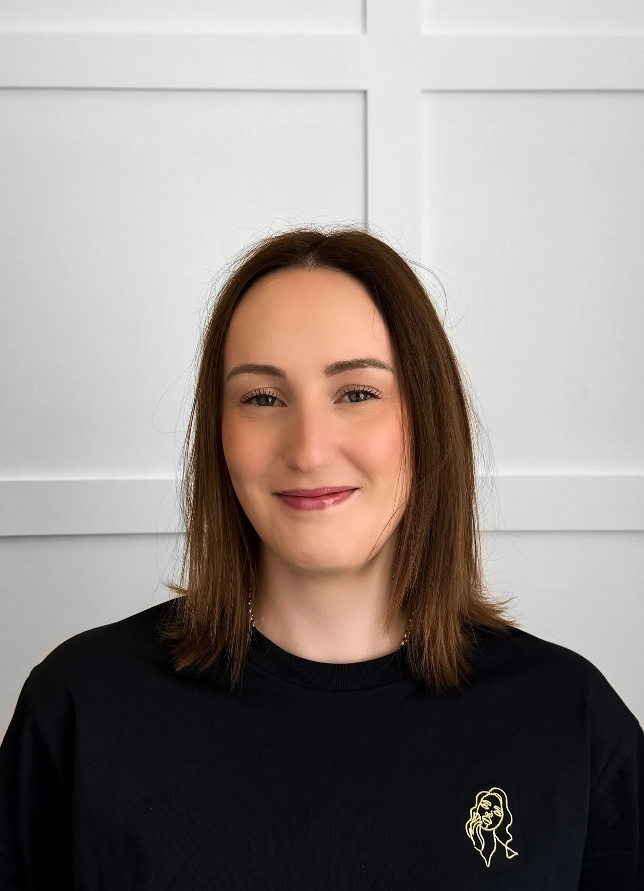 A woman with shoulder-length brown hair, wearing a black t-shirt with a small line art drawing of a woman's face on the chest, standing in front of a white paneled wall, smiling softly.