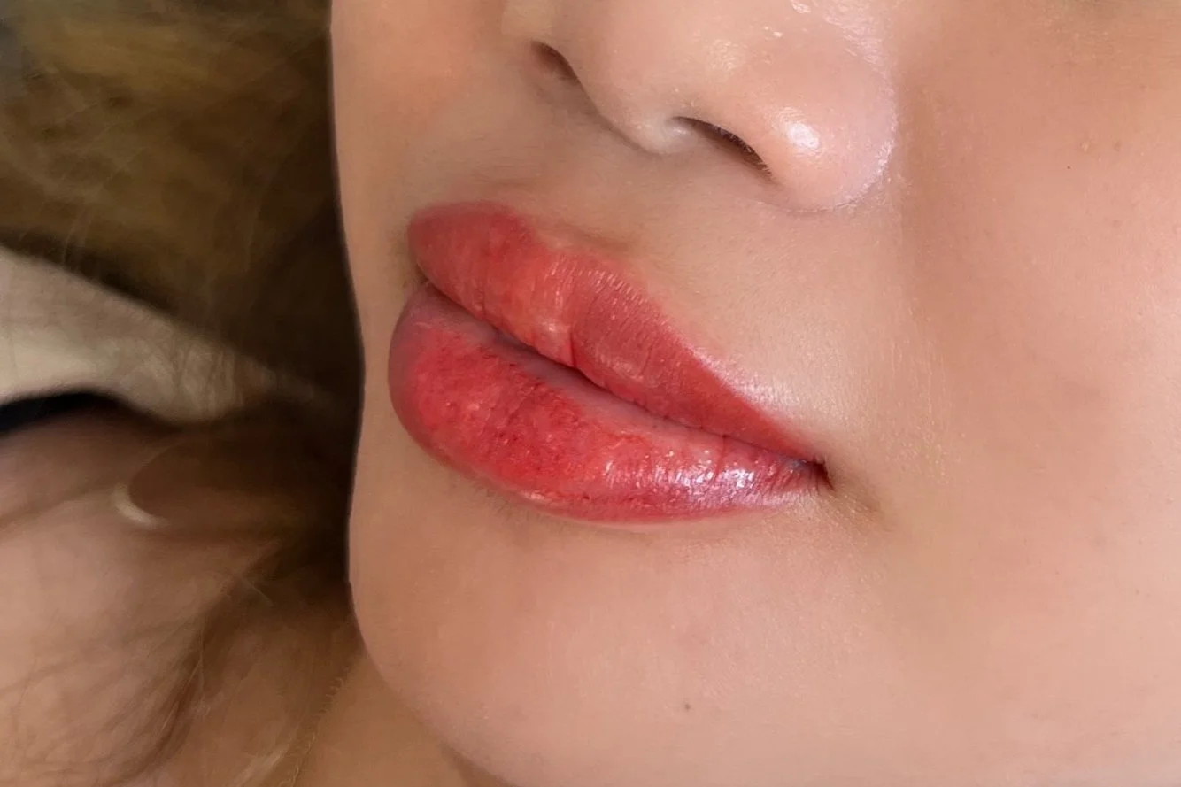Close-up of a person's lips with bright red lipstick, part of their nose and cheek, and some brown hair visible on the side.